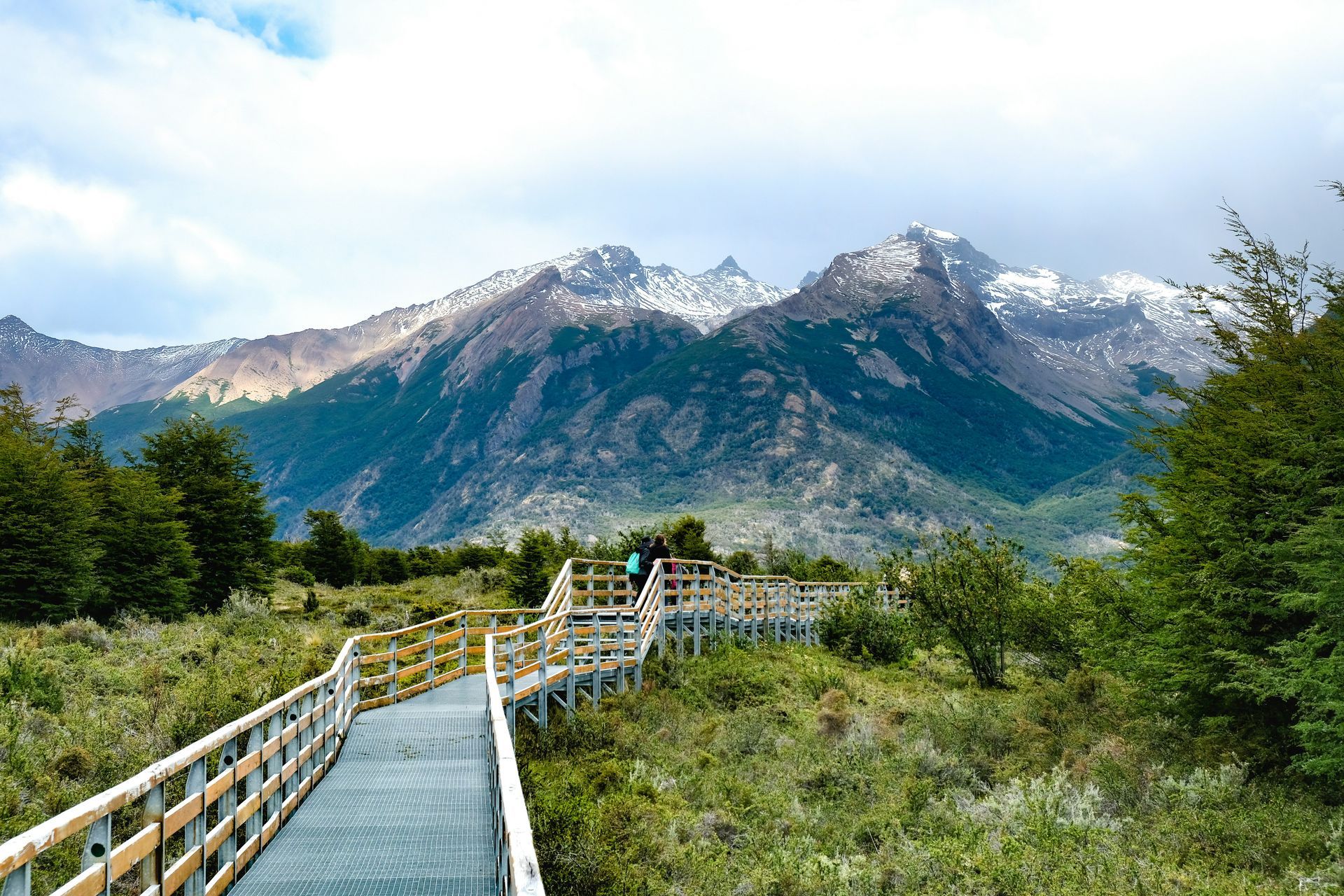 Wooden walkway through green landscape with snow-capped mountains at Parque Nacional Los Glaciares in El Calafate, Argentina.