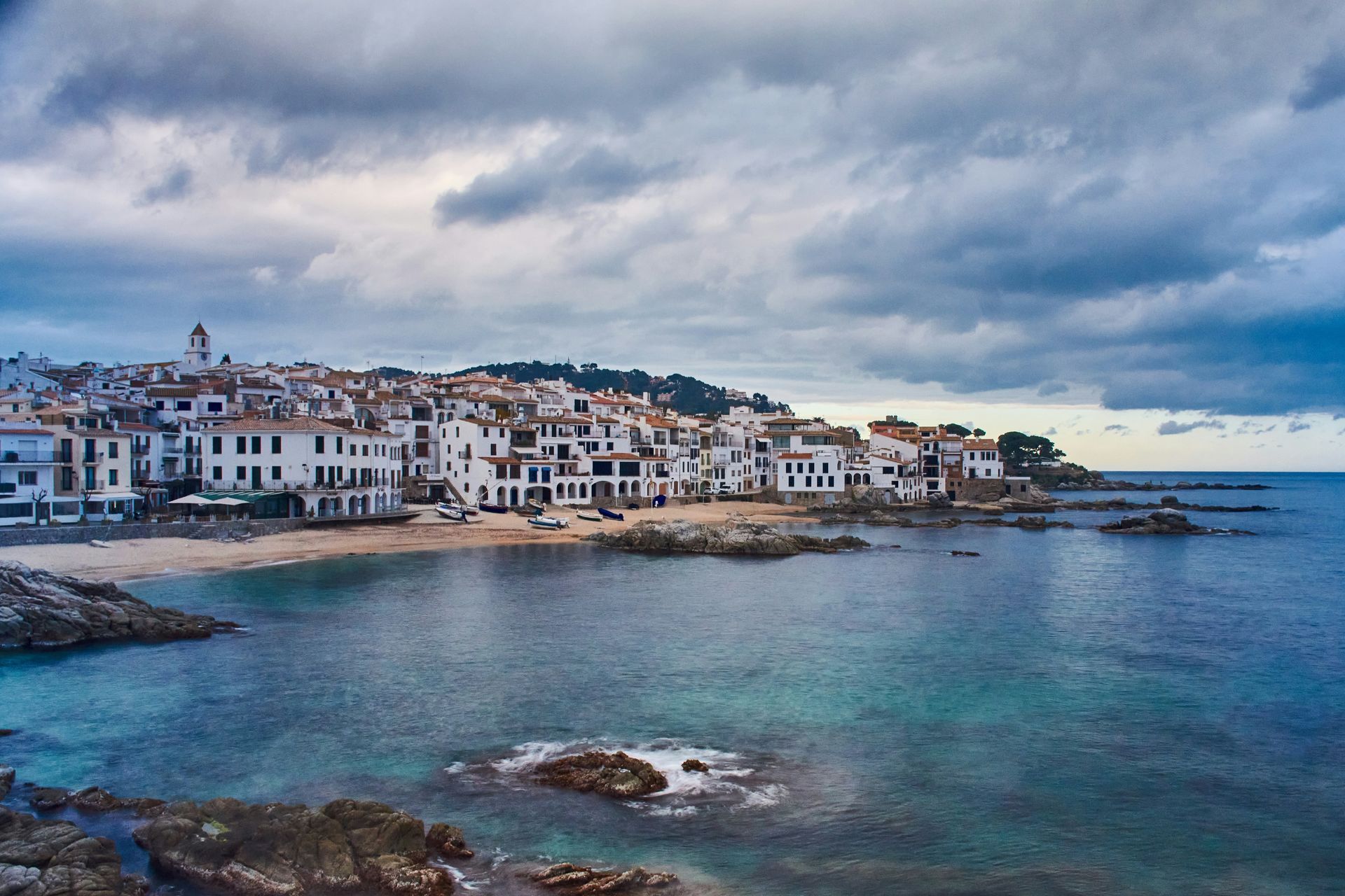 Coastal town with white buildings, sandy beach, and blue water under cloudy sky on Costa Brava, Spain.