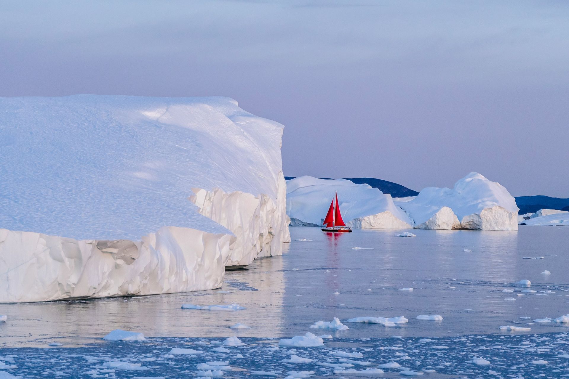 Red sailboat navigating icy Arctic waters near large icebergs in Disko Bay, Greenland.