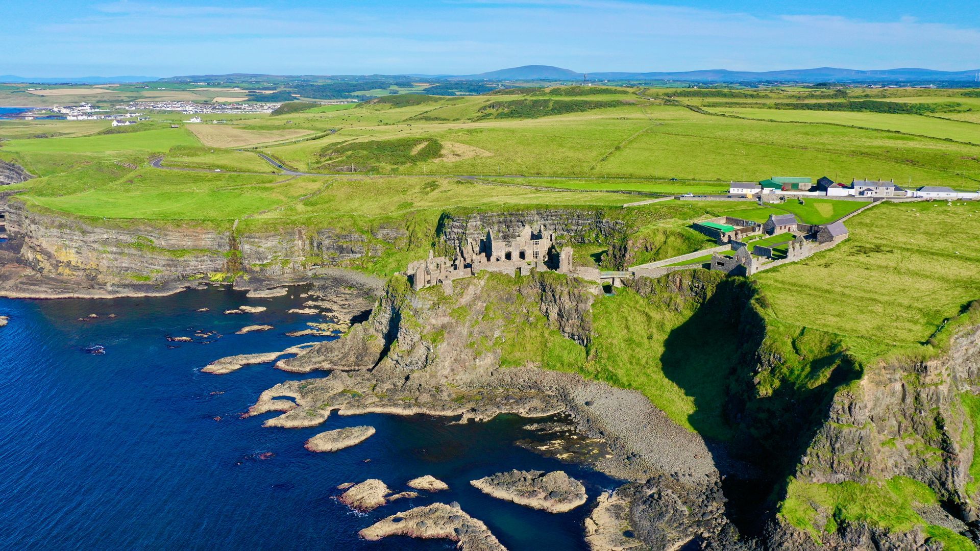 Ruins of Dunluce Castle on a green cliff overlooking the blue ocean under a bright blue sky in Northern Ireland.