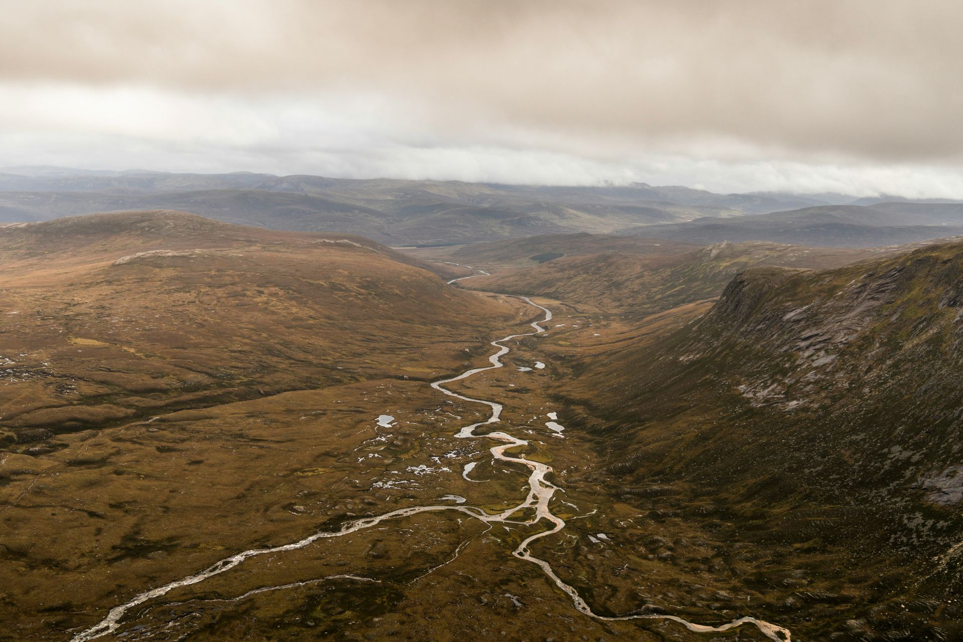 Valley landscape with winding river, brown terrain, under cloudy sky in Cairngorms National Park, Scotland.