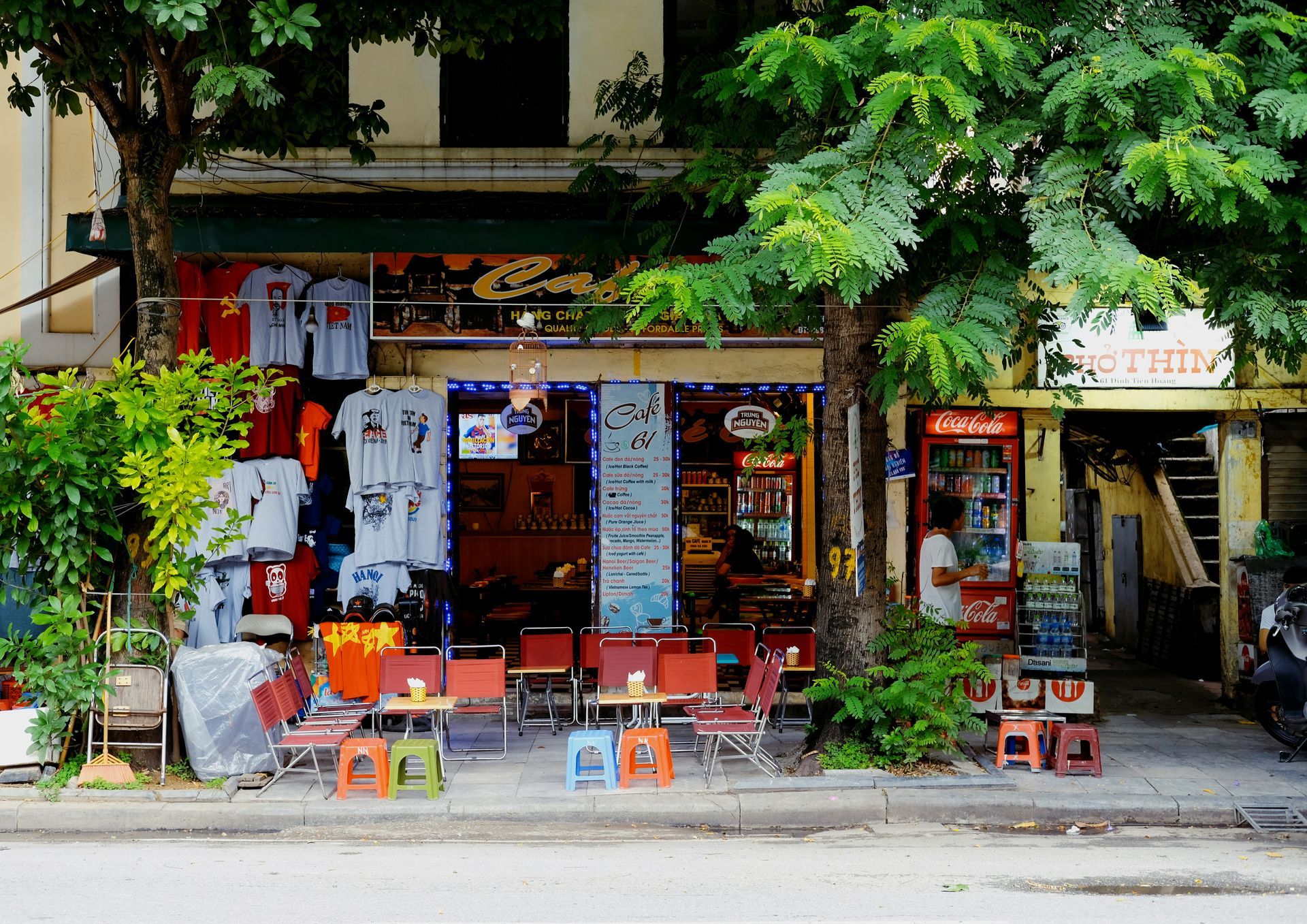 Cafe storefront with tables, chairs, t-shirts, and leafy trees in Hanoi, Vietnam.