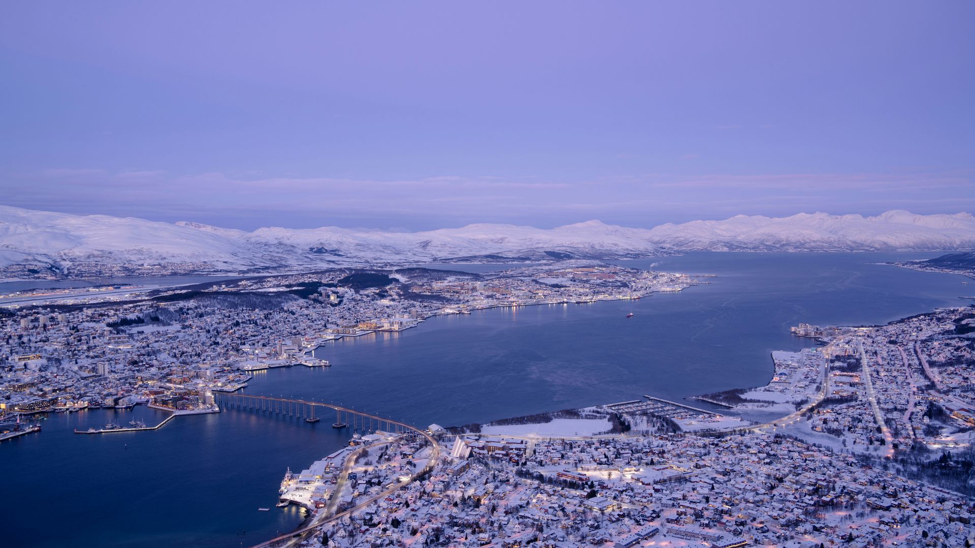Snow-covered cityscape with a bridge over a dark blue waterway under a dusky sky in Tromsø, Norway.