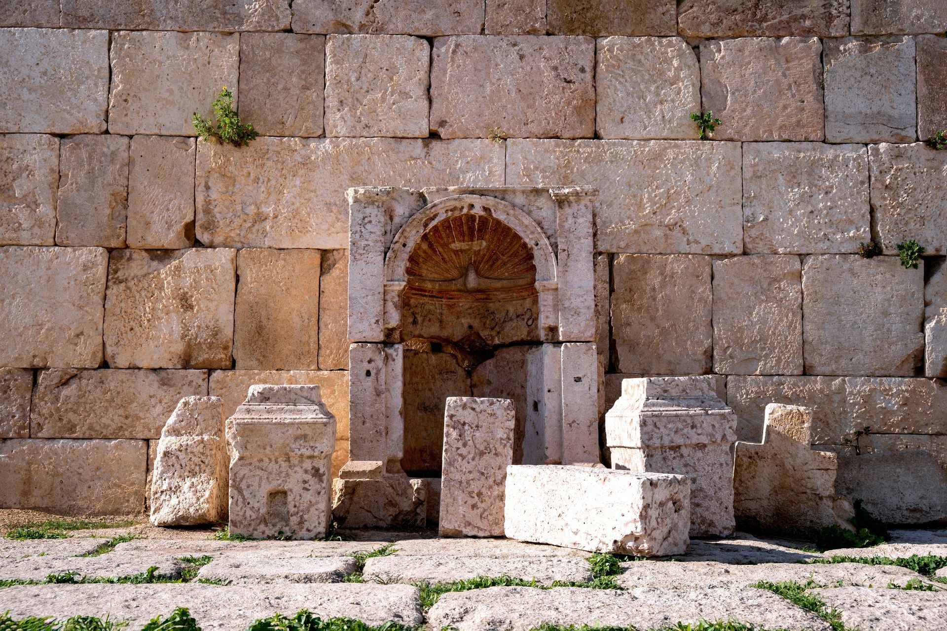 Stone ruins of an archway within a larger stone wall, with fragments in front in Jerash, Jordan.