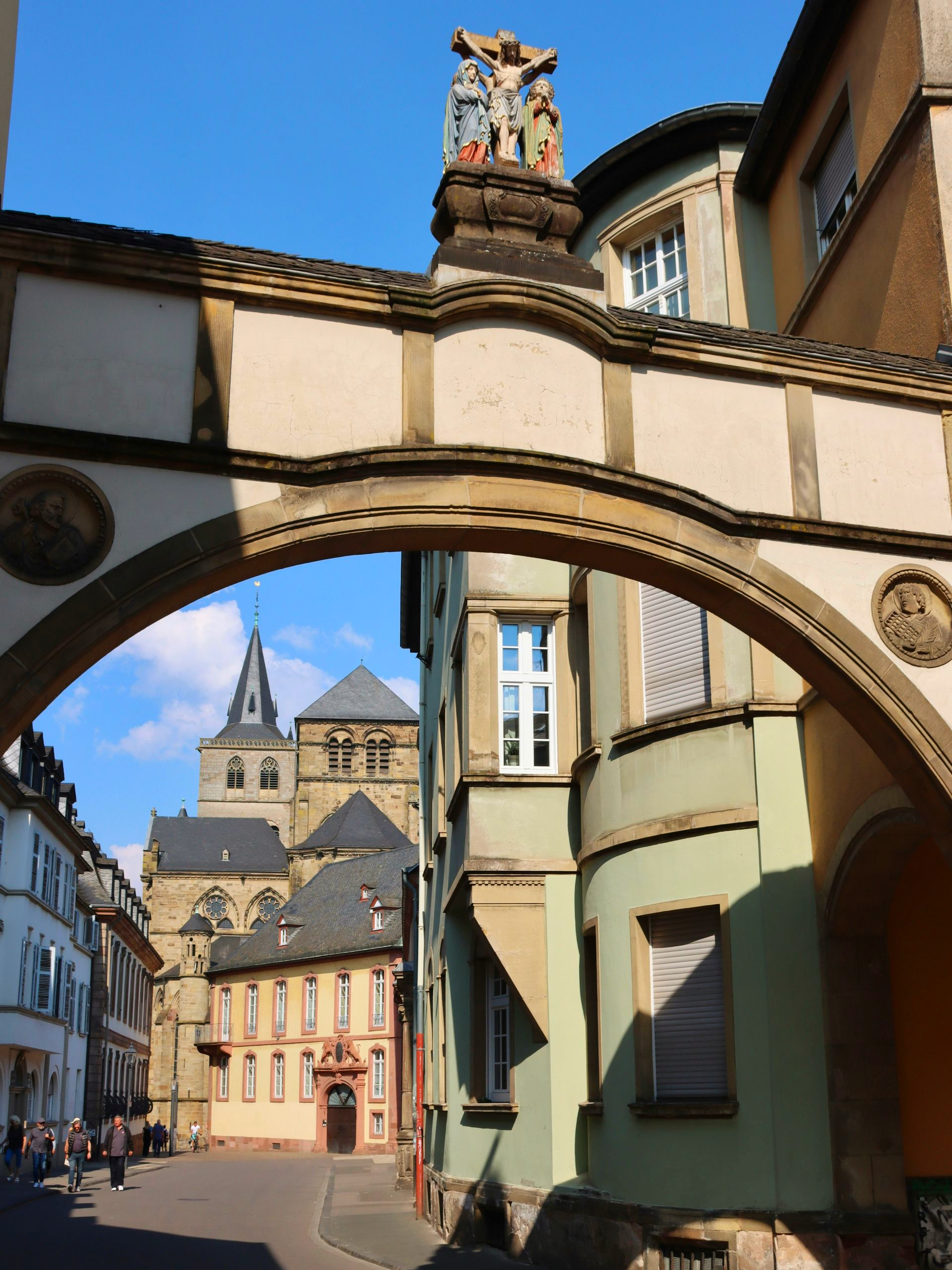 Archway over a street in Trier, Germany, with buildings, a church, and a bright sky.