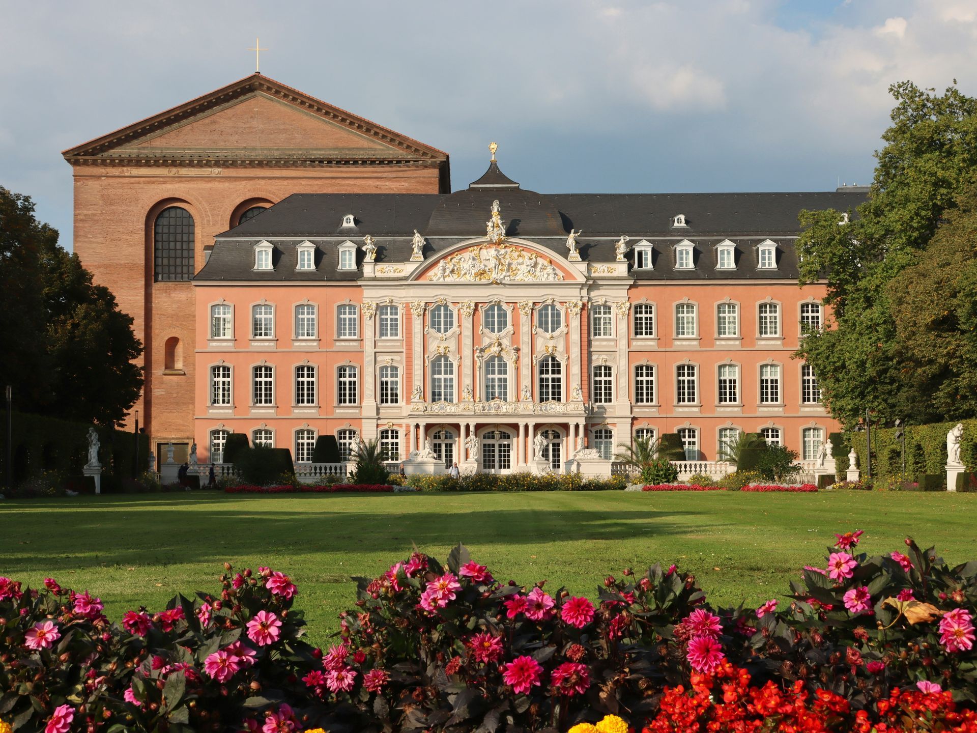 Pink palace in Trier, Germany with central ornate section, red-brown brick wing on left, green lawn, red and pink flowers.