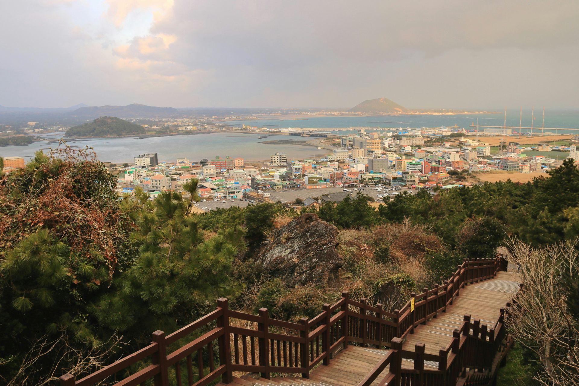 View from a hilltop path overlooking a coastal city, harbor, and distant islands on Jeju Island, South Korea.