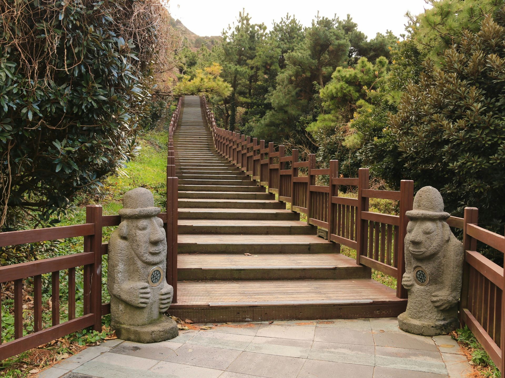 Stone stairs with wooden railing lead up a hill, flanked by statues and trees on Jeju Island, South Korea.
