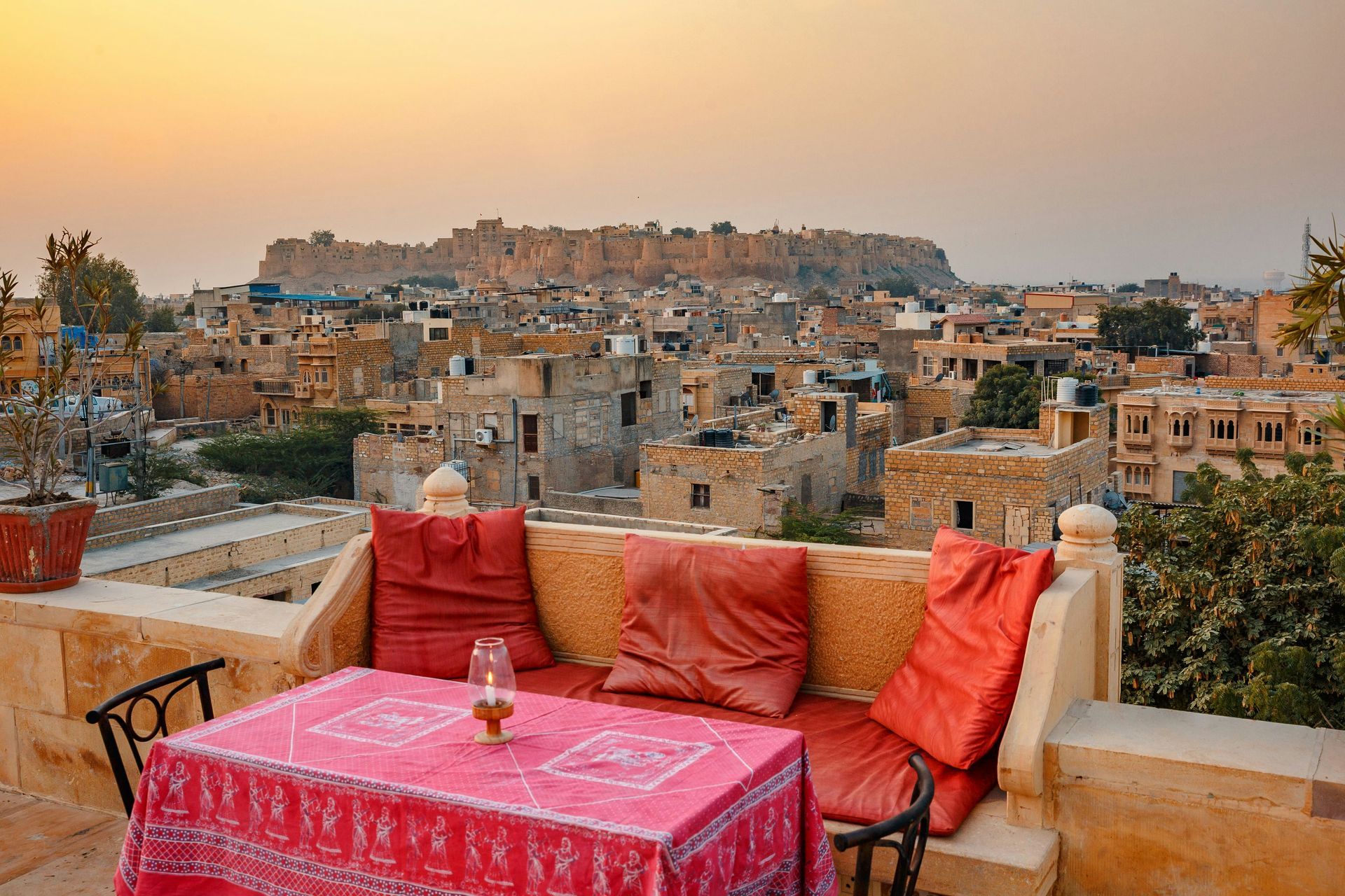 Rooftop cafe in Jaisalmer, India, with a view of the sandstone city at sunset. Red cushions and table, buildings.
