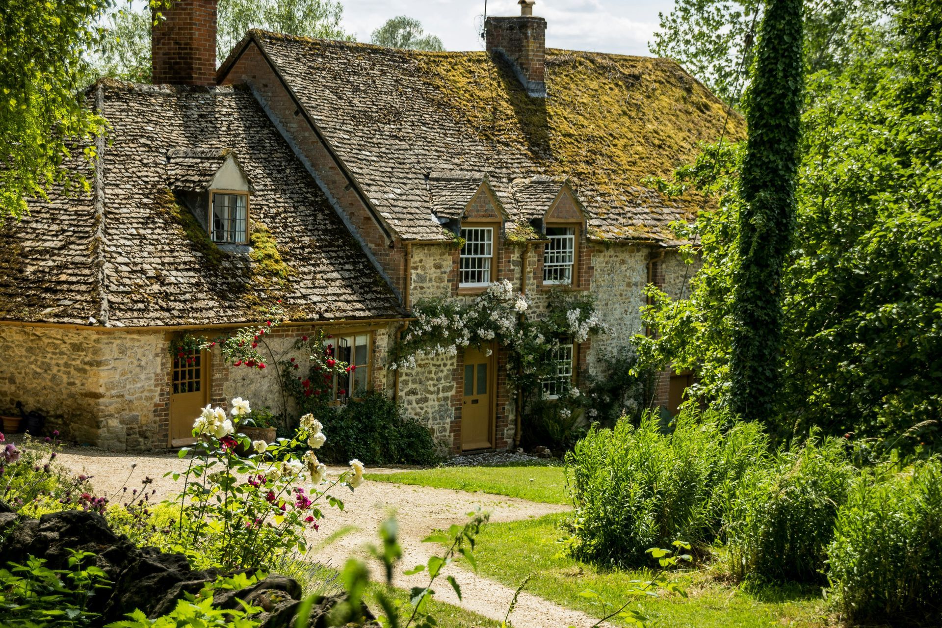 Stone cottage with a mossy roof, surrounded by green foliage and a winding gravel path in the Cotswolds, England.