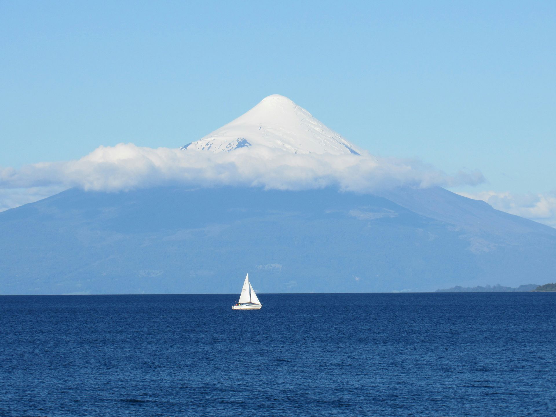 Sailboat on blue water with a snow-capped volcano in the background. Bright blue sky in Los Lagos, Chile.
