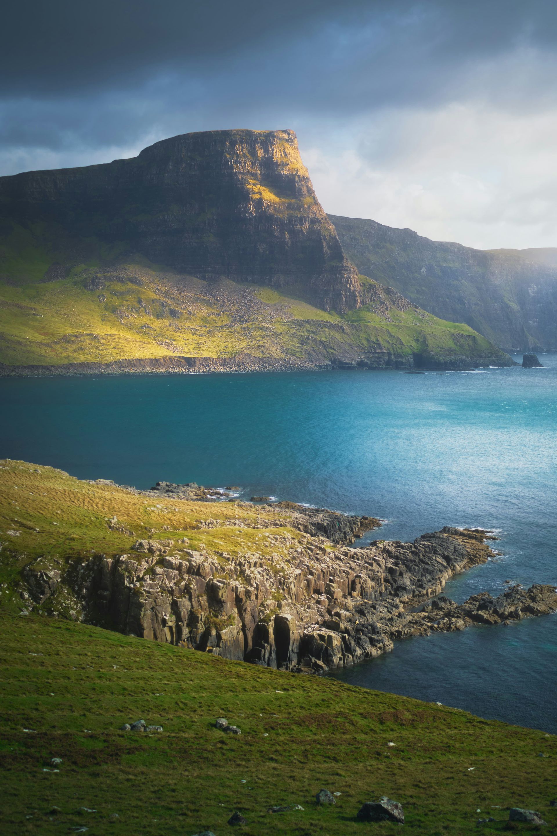 Cliffs under a mountain with turquoise water. Green grass in foreground, dramatic sky overhead on the Isle of Skye, Scotland.