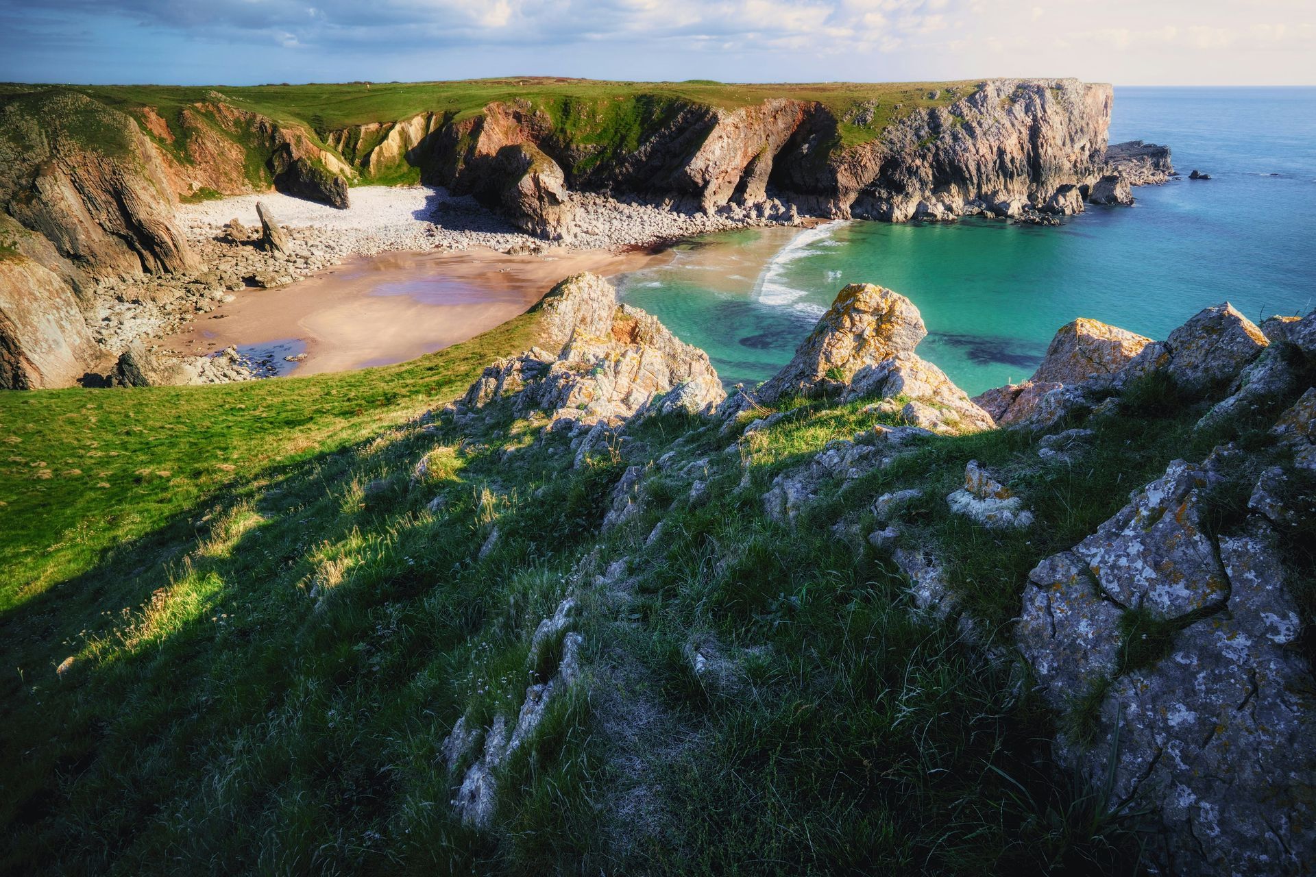 Green cliffside overlooking a turquoise bay with rocky beaches and a blue sky in Pembrokeshire, Wales.