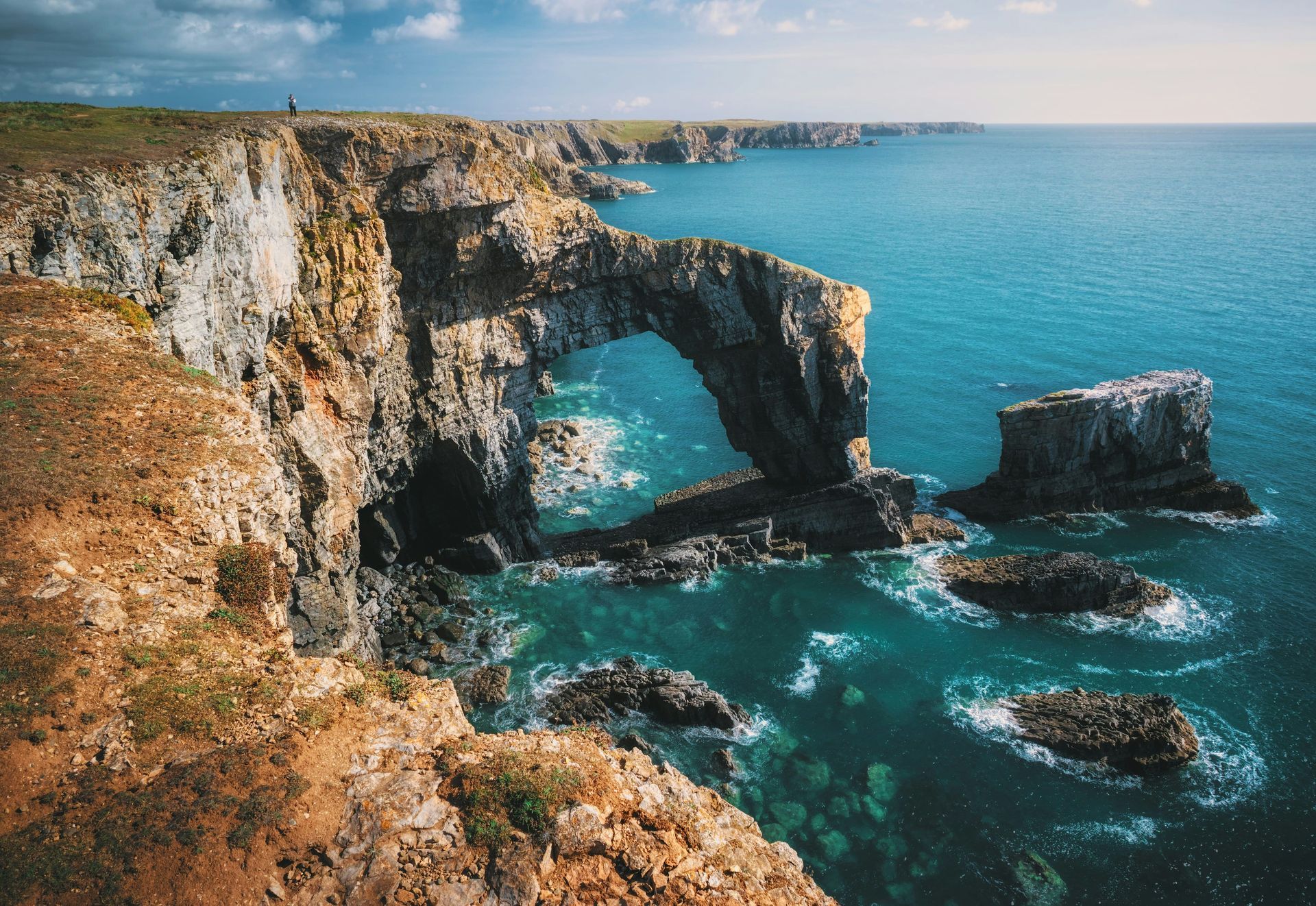 Coastal arch of rock carved by ocean waves; turquoise water, blue sky in Pembrokeshire, Wales.