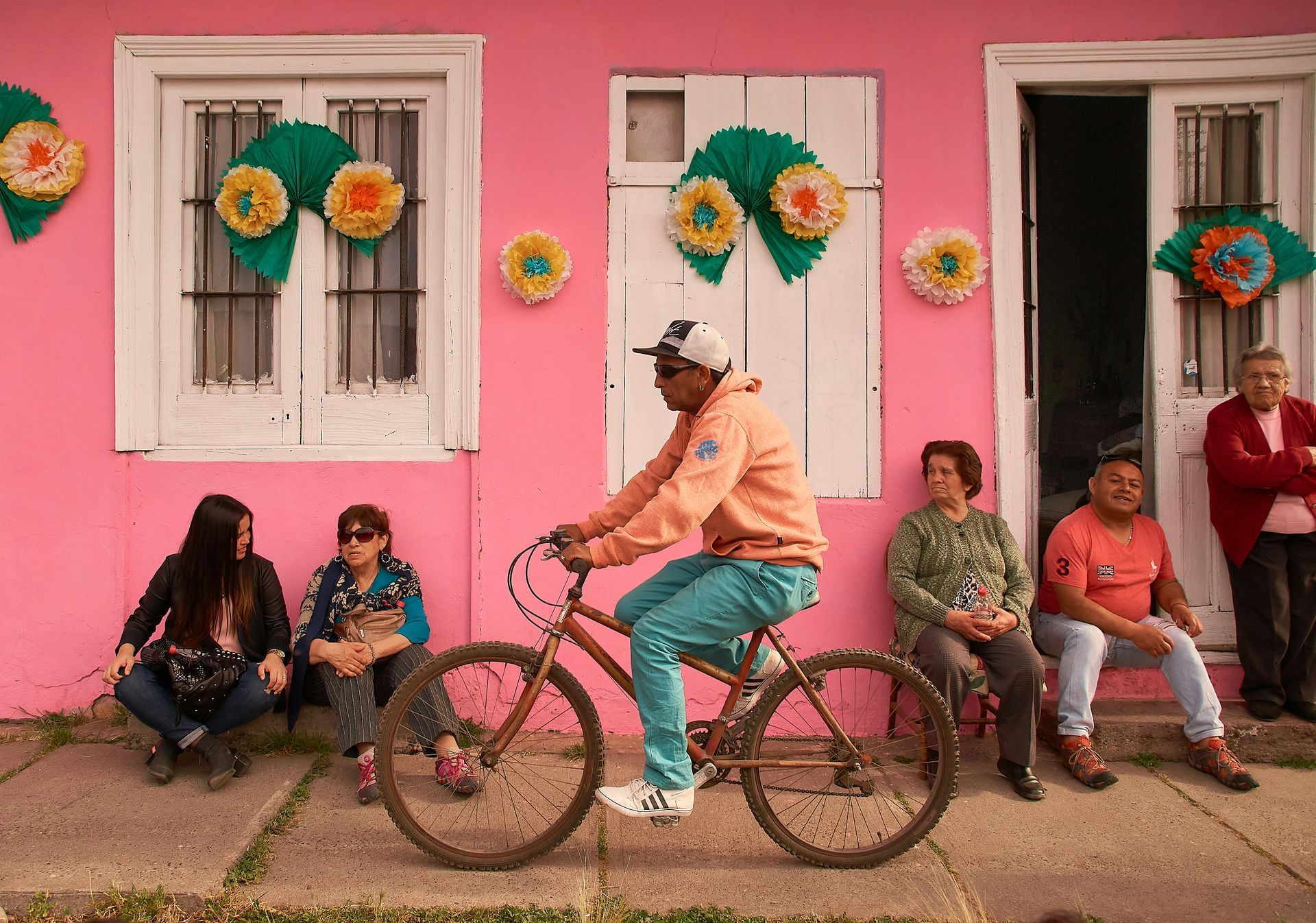 A person riding a bicycle past people sitting near a pink house decorated with colorful paper flowers in Chile.