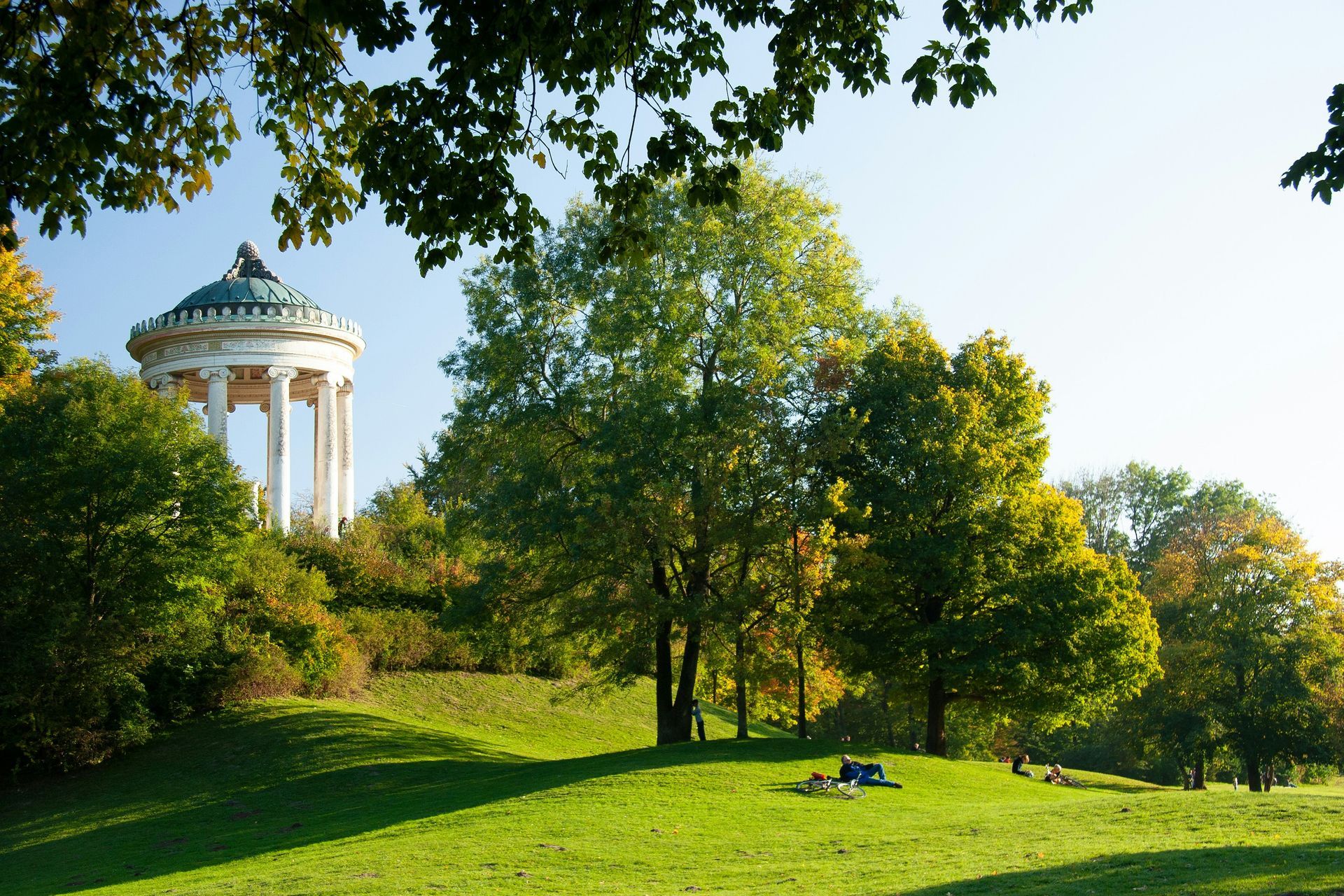 A green park with a gazebo on a hill, trees, and people relaxing on the grass at the Englischer Garten, Munich.