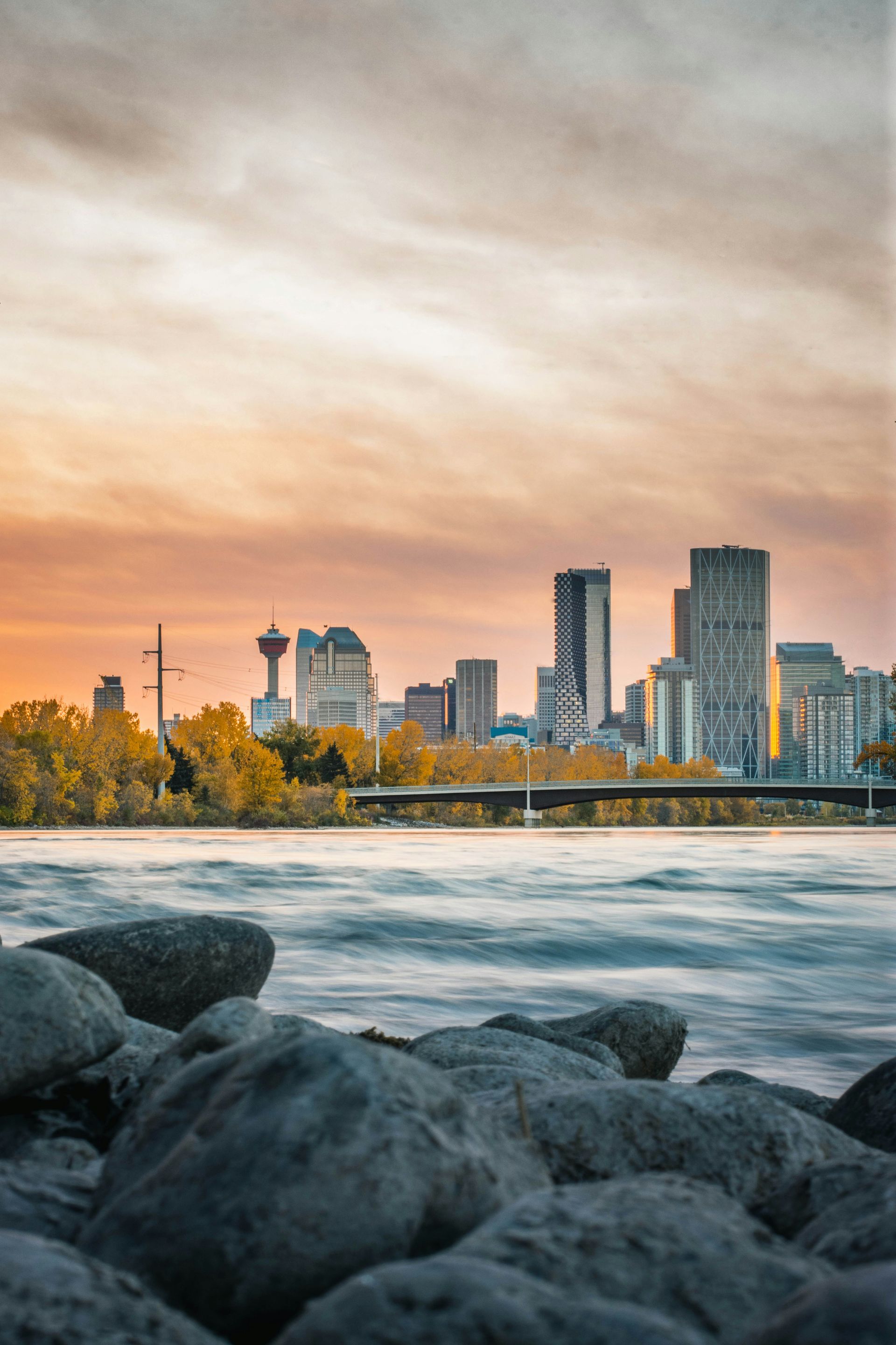 Calgary's skyline at sunset with an orange-toned sky, viewed over a river with rocks in the foreground.