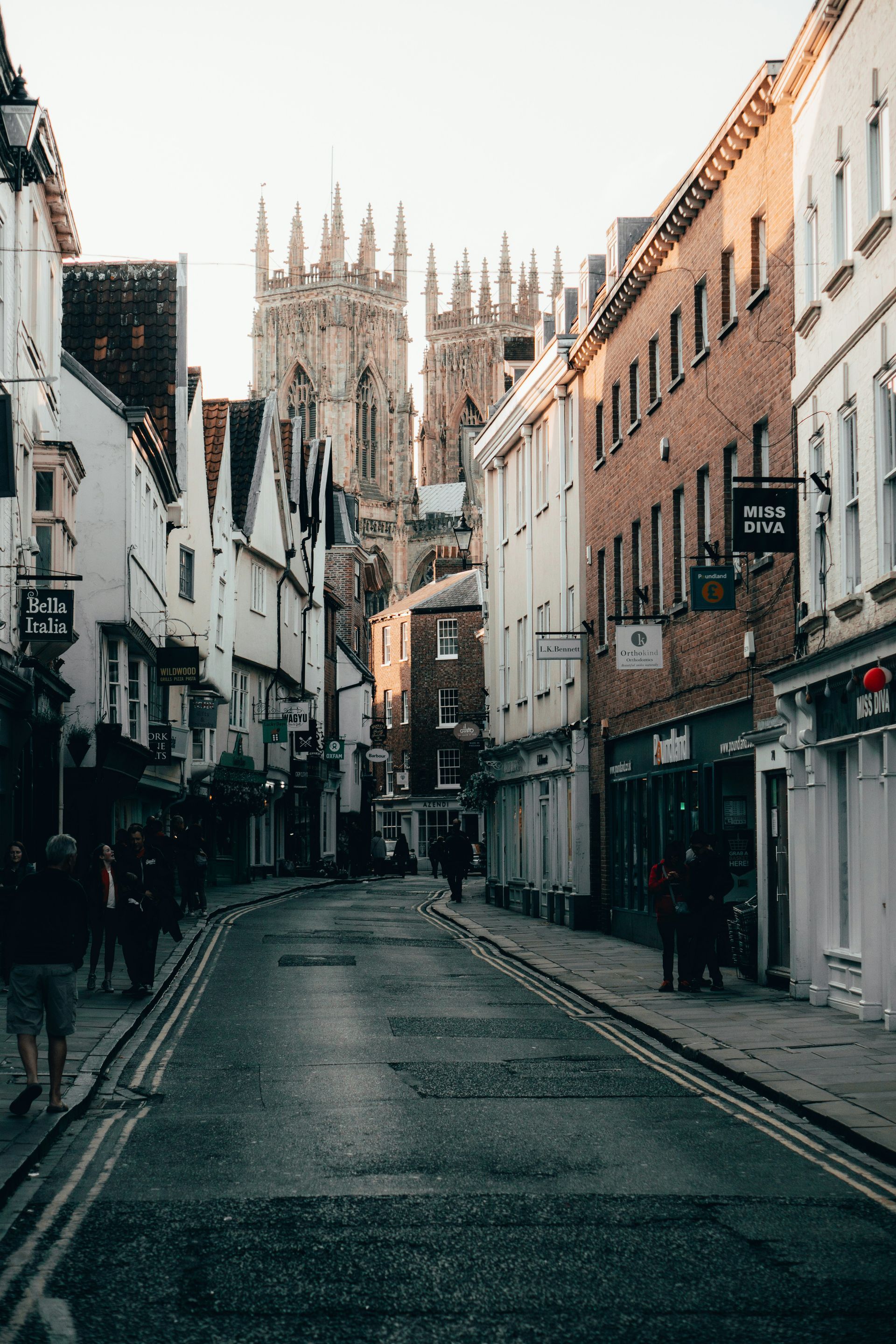 Cobblestone street in York, UK, leads toward a towering cathedral; lined with shops and buildings.