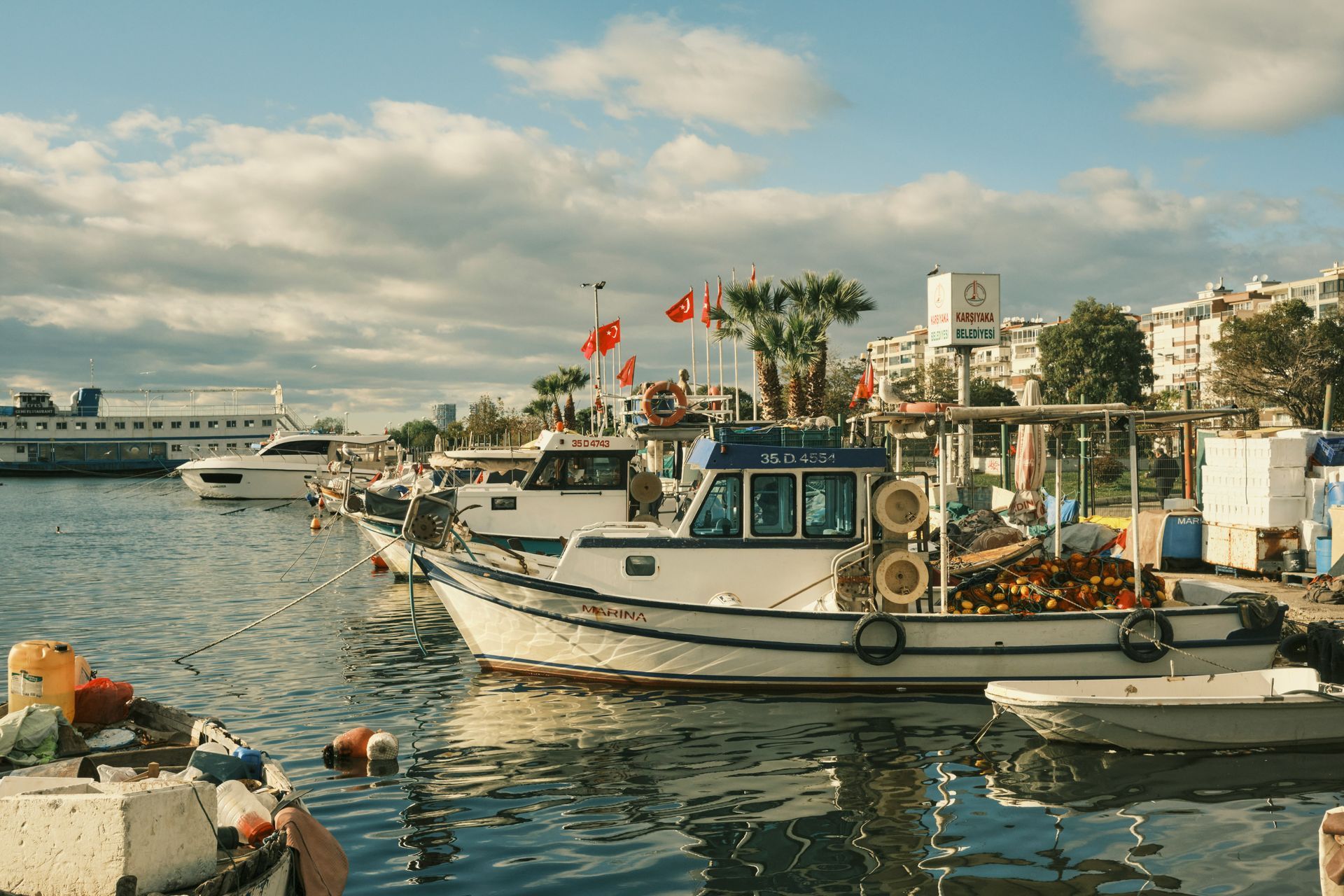 Boats docked at a harbor under a partly cloudy sky. Red flags in İzmir, Turkey.