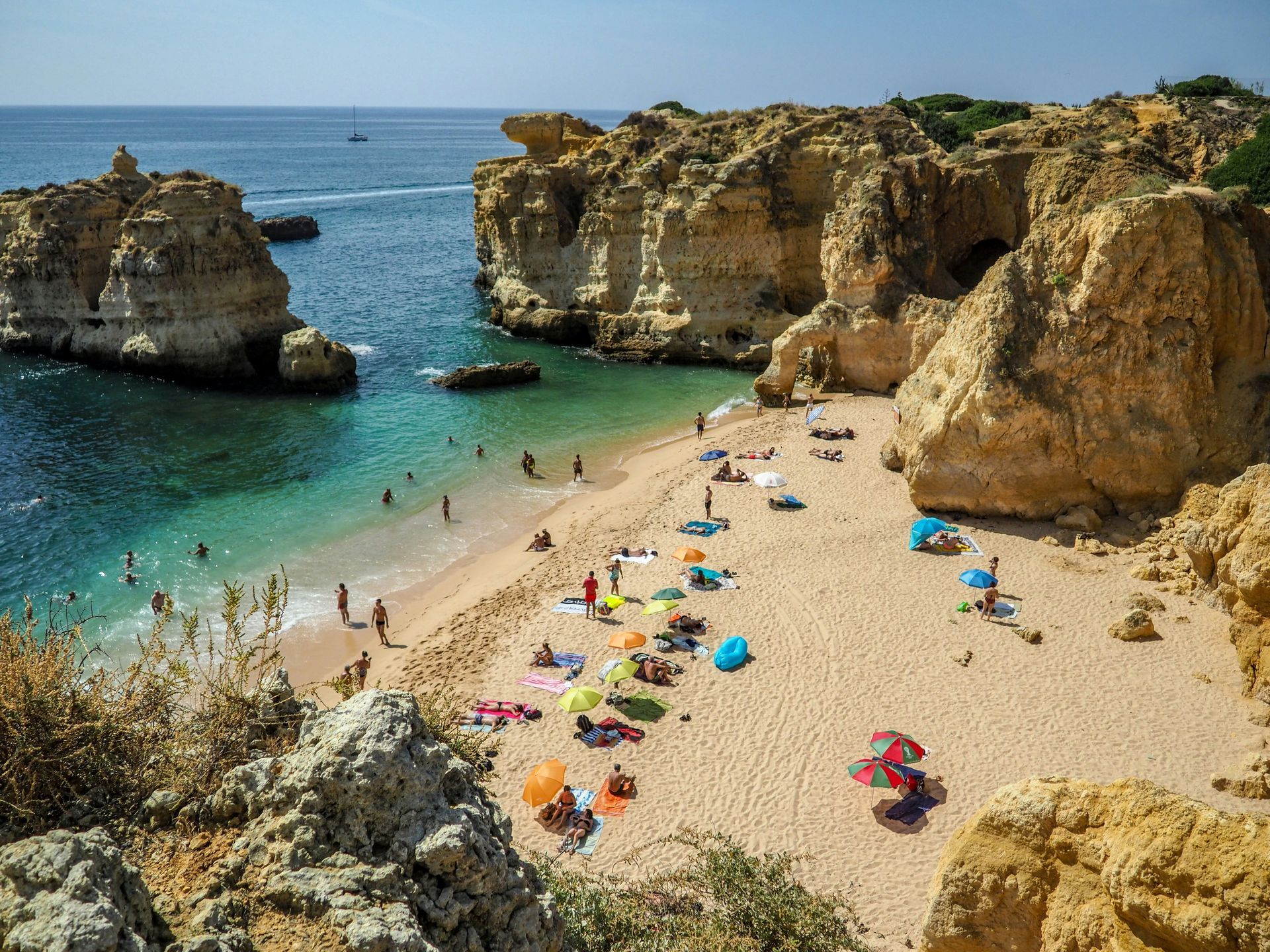 Beach with turquoise water, rocky cliffs, and people relaxing under colorful umbrellas in The Algarve, Portugal.
