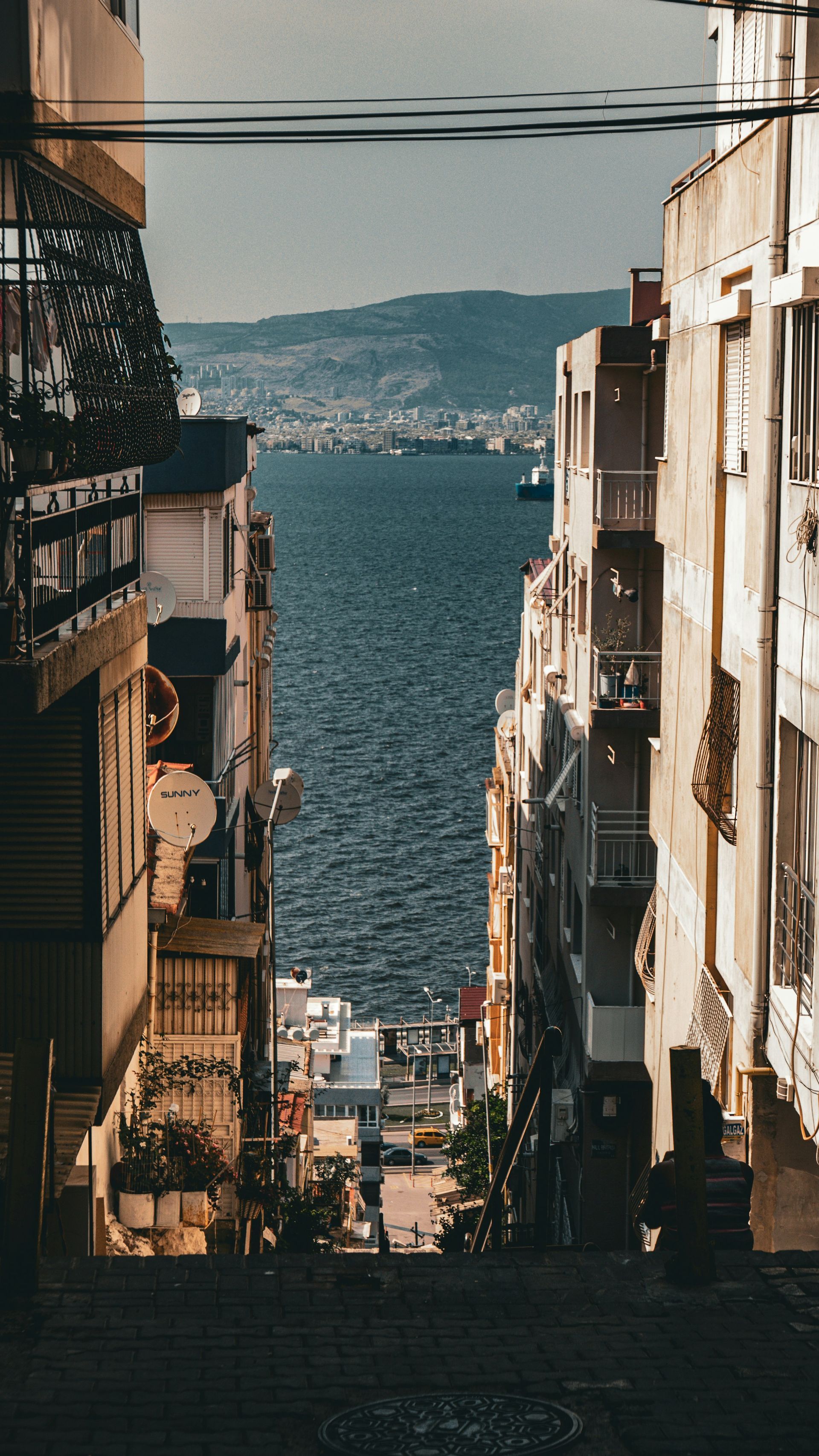 A narrow street of buildings leads to a view of the sea and distant hills in İzmir, Turkey.