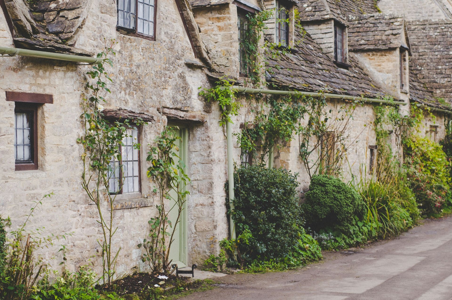 Cotswold cottages with stone walls, ivy, and a green door, along a narrow road in England.