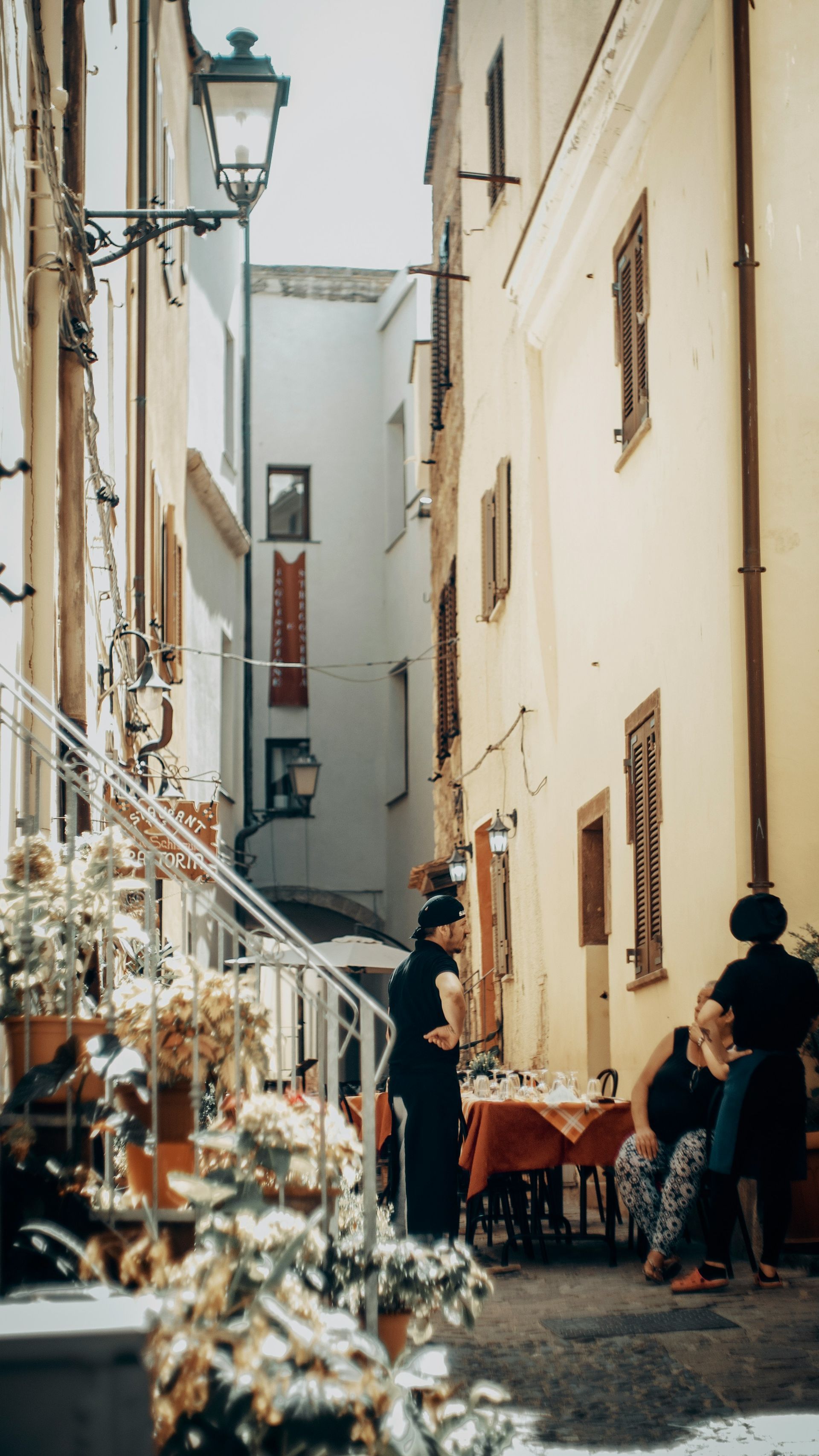 A group of people are sitting at tables in a narrow alleyway between two buildings in Sardinia, Italy.
