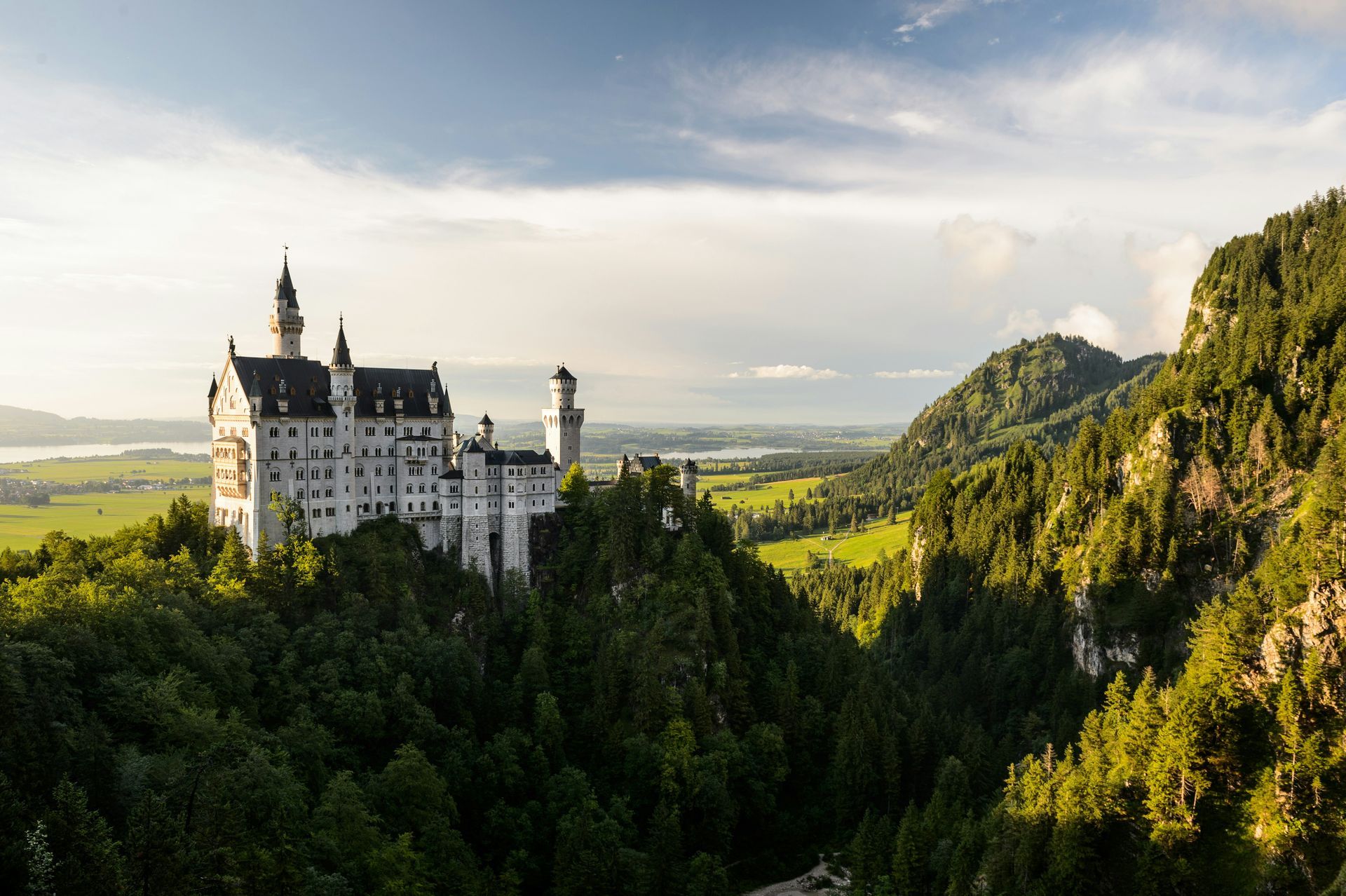 Neuschwanstein Castle atop a green, forested mountain, under a partly cloudy sky.