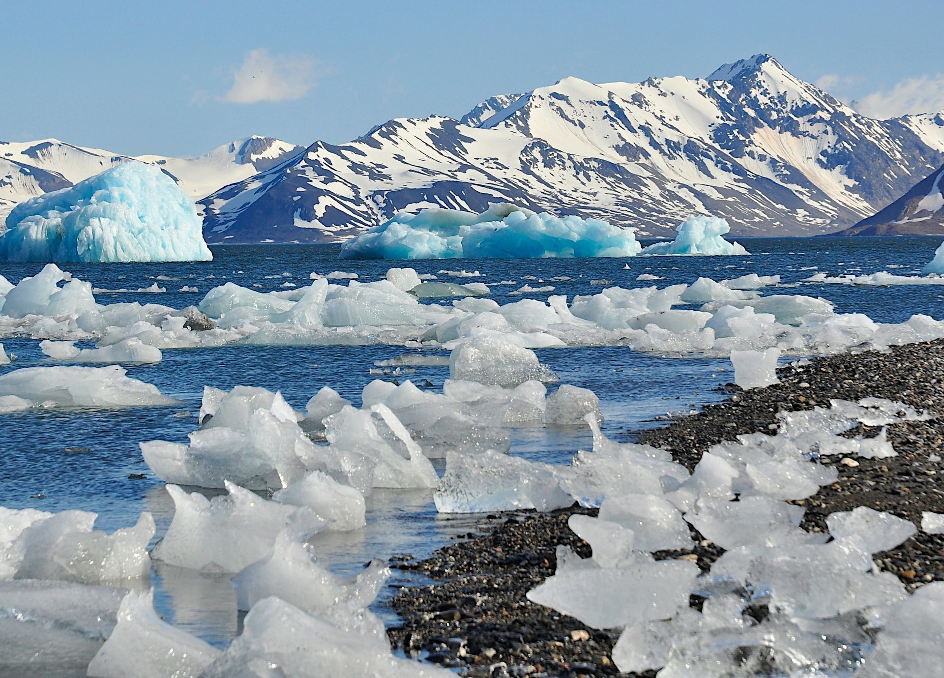 Icebergs and sea ice in a blue ocean with snowy mountains under a partly cloudy sky in Svalbard, Norway.