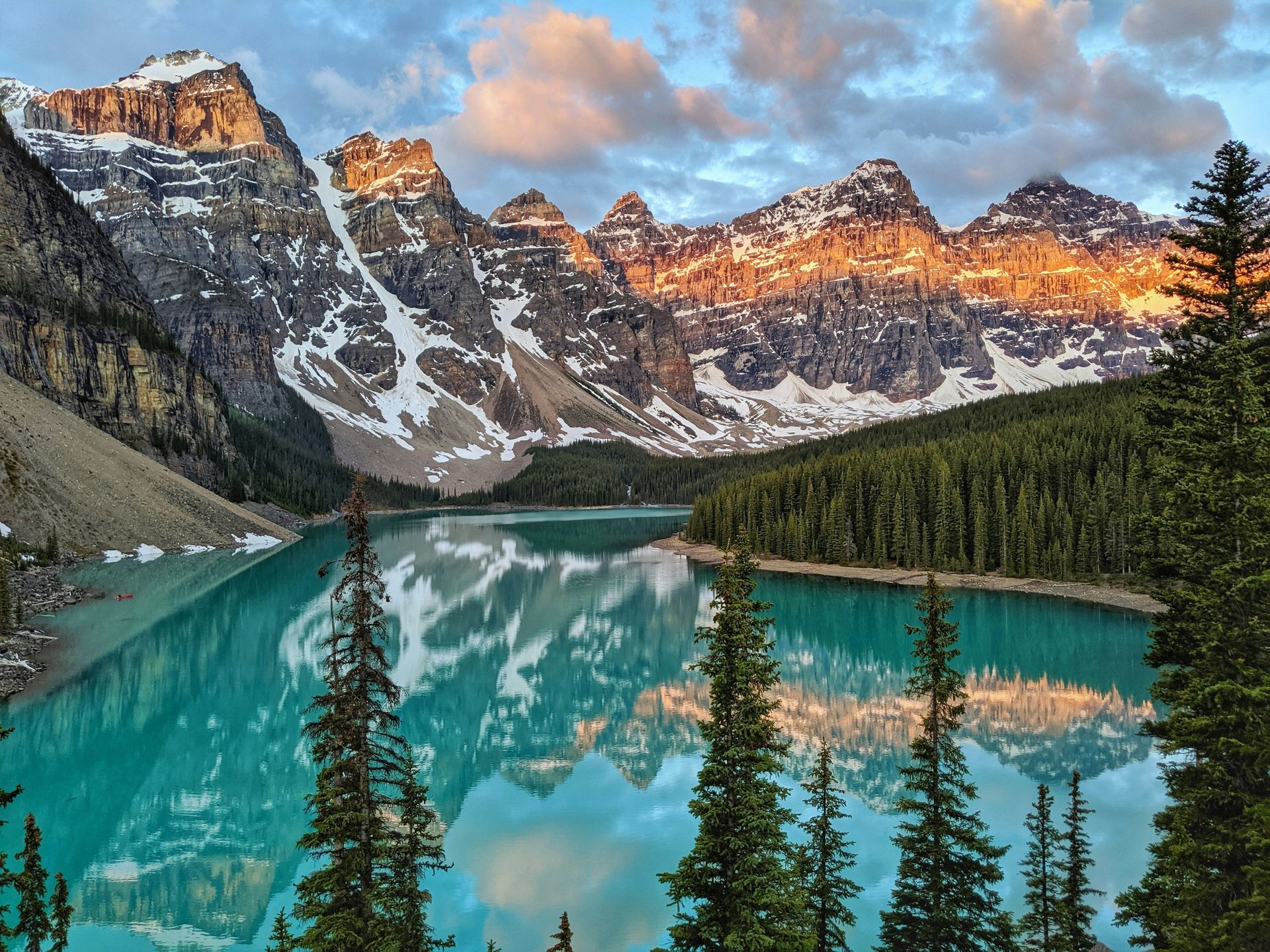 Turquoise Moraine Lake reflects snow-capped mountains lit by golden sunlight; evergreen trees frame the scene in Banff, Canada.