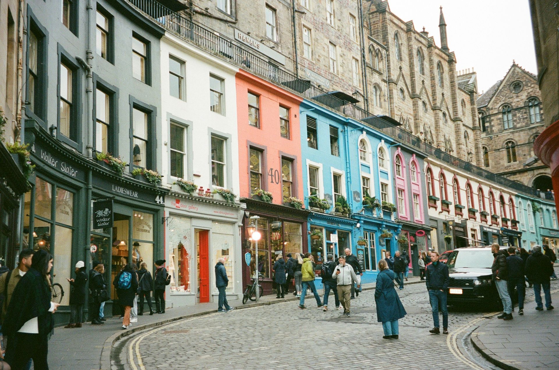 The colorful Victoria Street in Edinburgh is lined with vibrant painted shops and pedestrians.