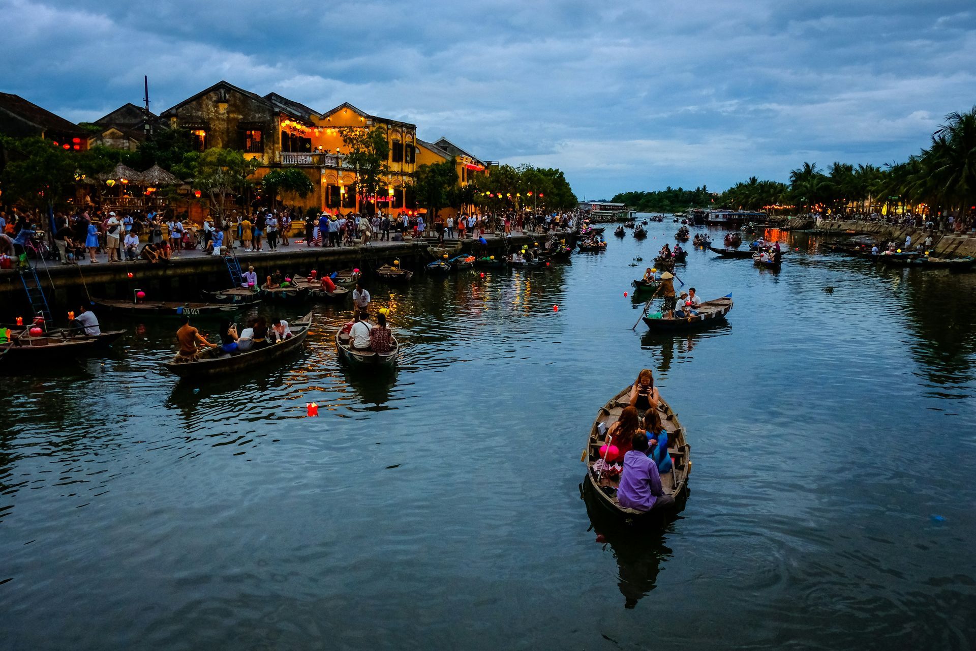 Boats on a river, people in buildings along the shore at dusk in Hoi An, Vietnam.
