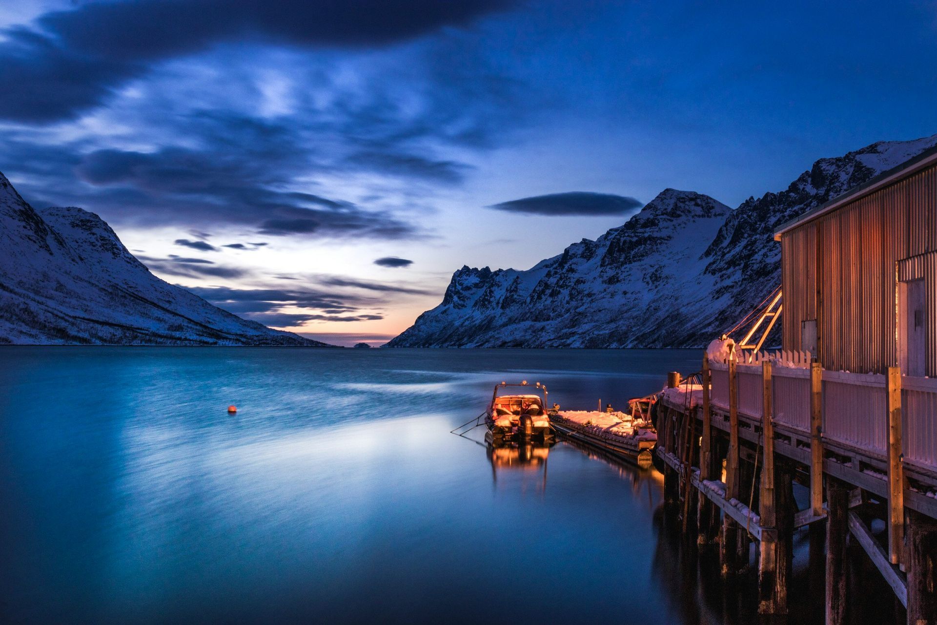 Snowy mountains frame a calm blue fjord at twilight, a wooden structure with pier on the right in Tromsø, Norway.