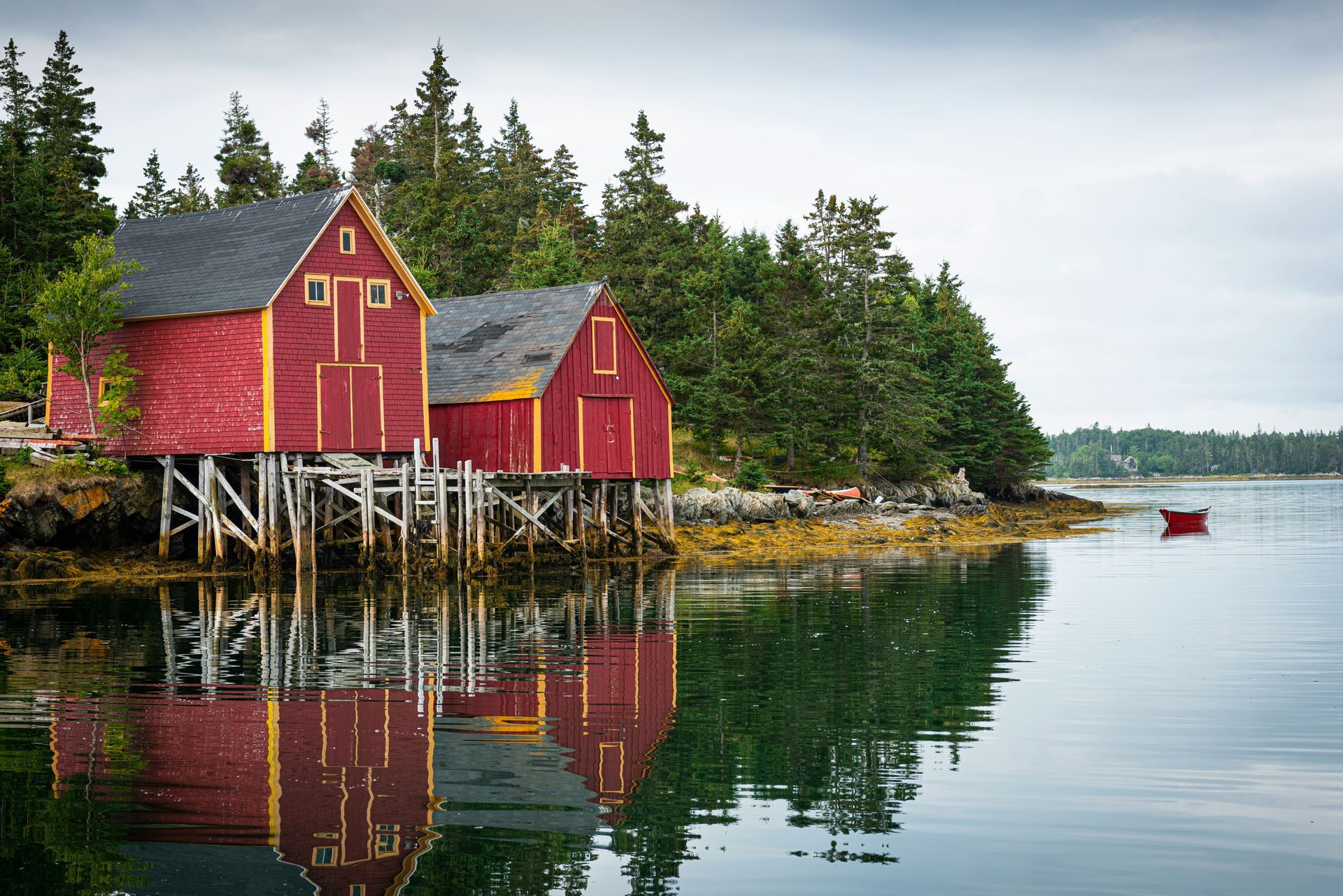 Two red wooden buildings on stilts reflected in calm water, trees in the background, small red boat in Nova Scotia, Canada.