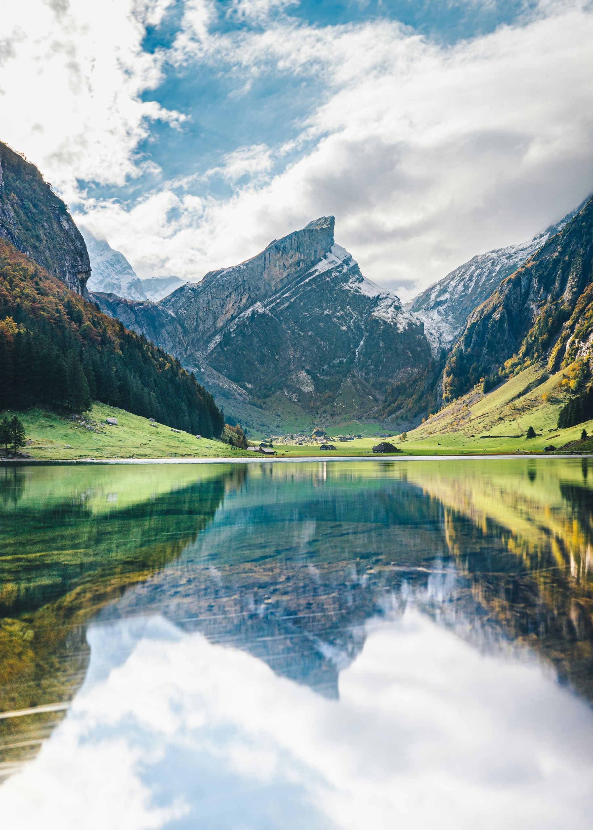 A reflective lake mirrors snow-capped mountains under a partly cloudy sky in Schwende, Switzerland.
