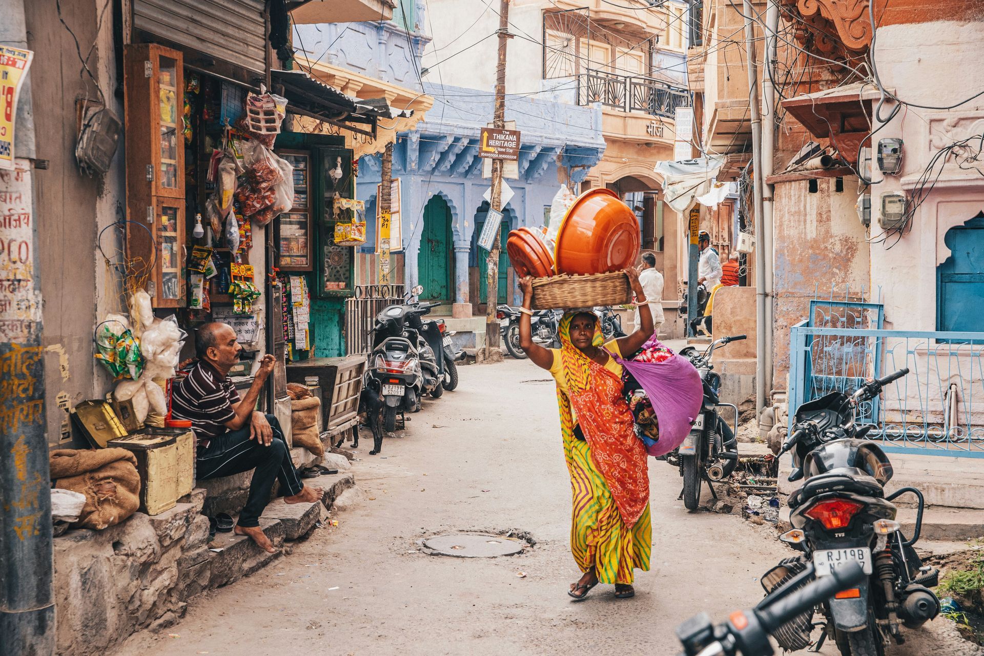 Woman carries bowls on her head through a bustling street in India, lined with shops, parked motorcycles, and colorful buildings.