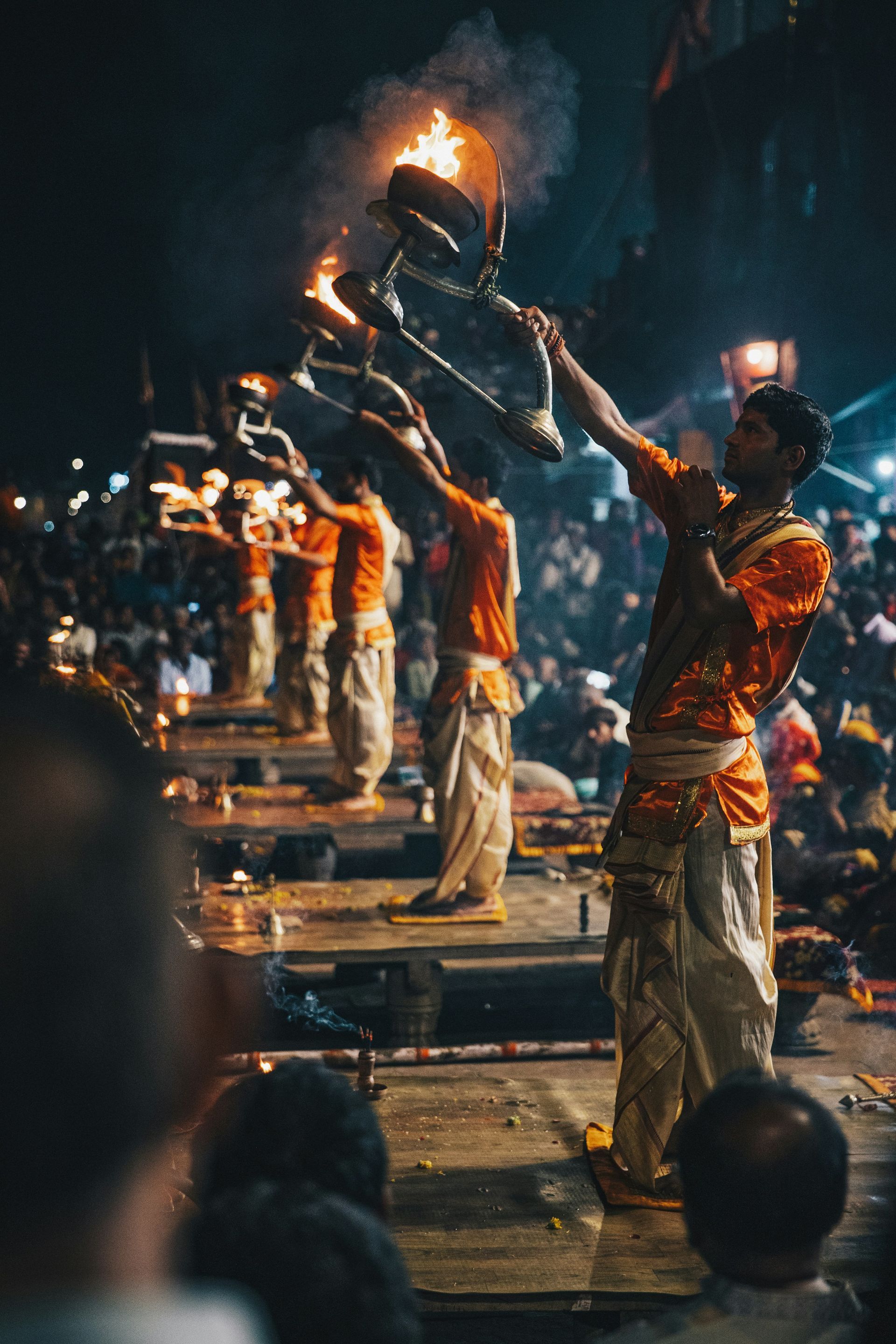 Men in orange and white clothing perform a nighttime Hindu fire ritual on steps by a river in Varanasi, India.