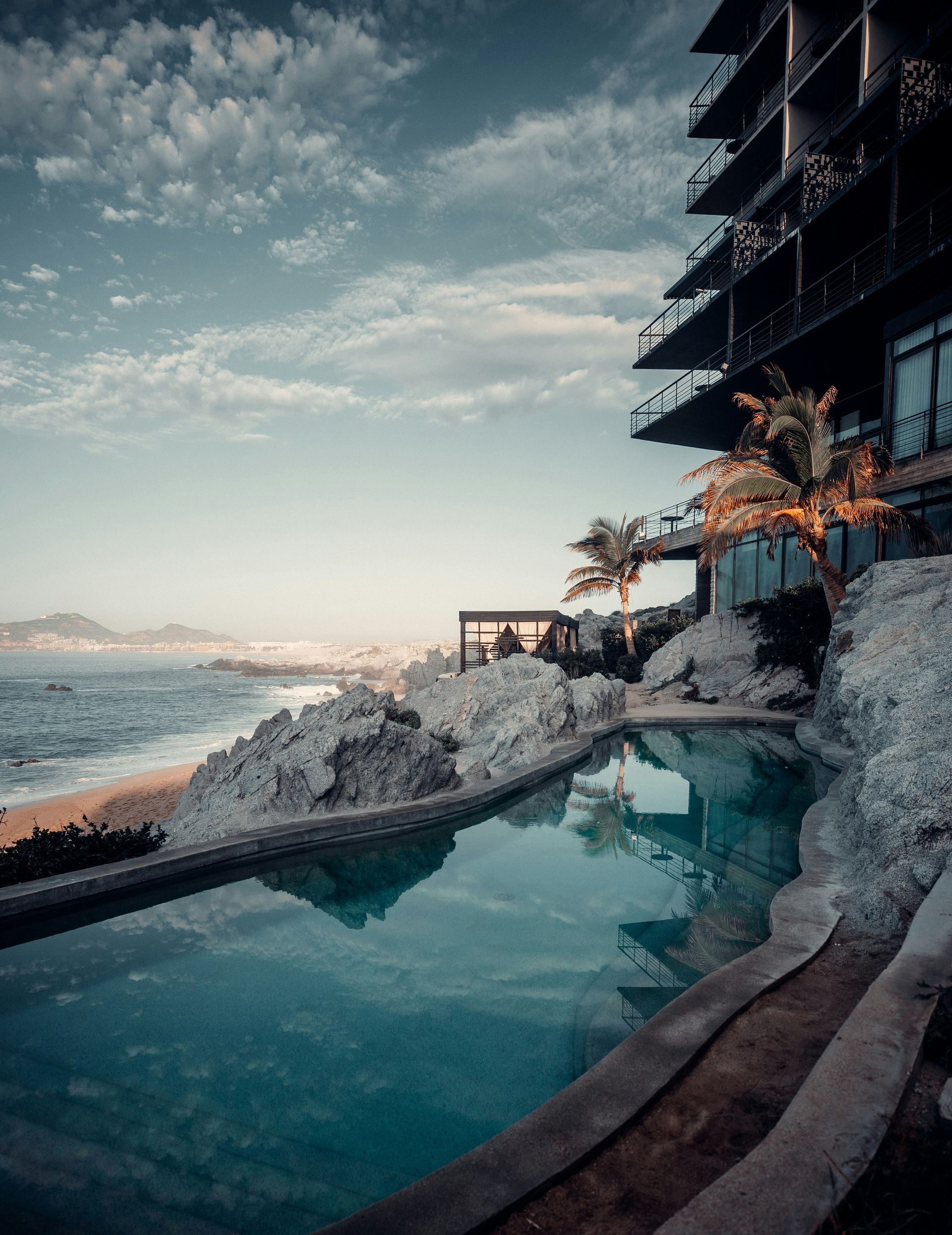 Infinity pool overlooking the ocean, next to a hotel on a cliff. Blue water and sky, palm trees in Cabo San Lucas, Mexico.
