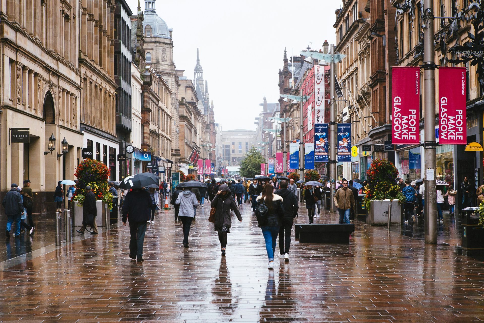 People walking on a wet city street in Glasgow, with shops, banners, and architecture.