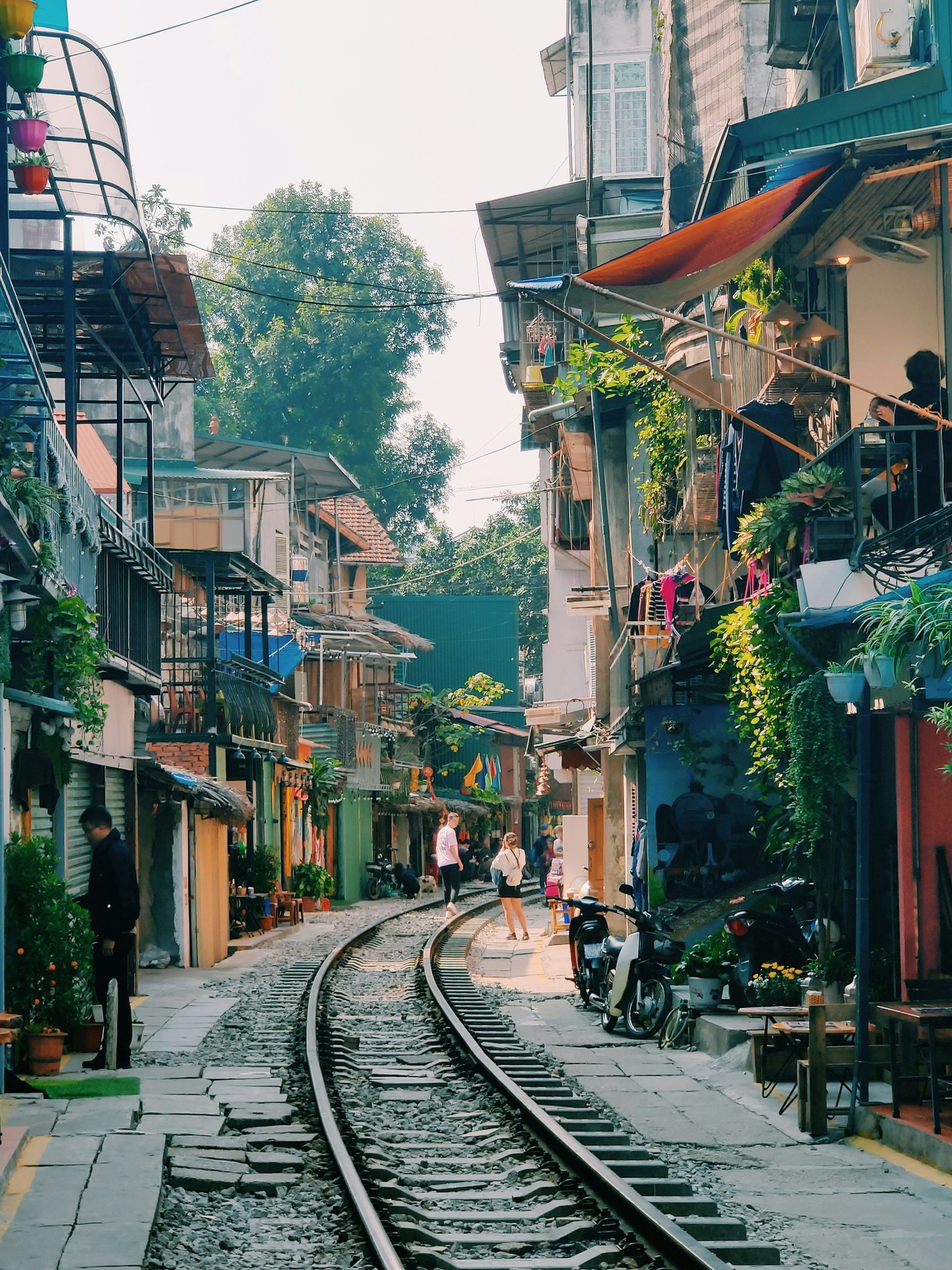 A narrow street with train tracks, lined with colorful buildings and greenery. People walk on the tracks in Hanoi, Vietnam.