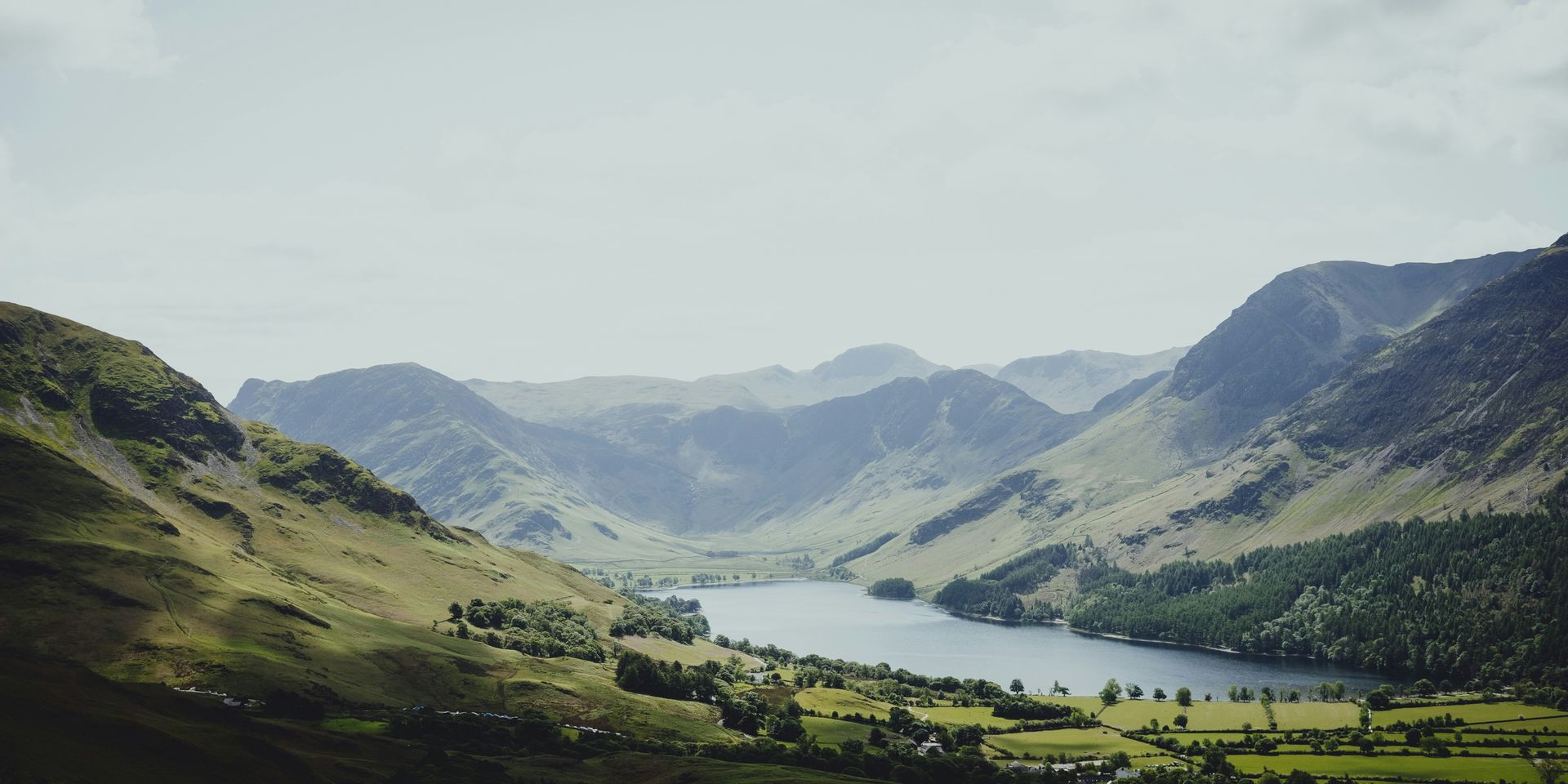 Mountain valley with a lake, flanked by green hills, under a hazy sky in the Lake District, England.