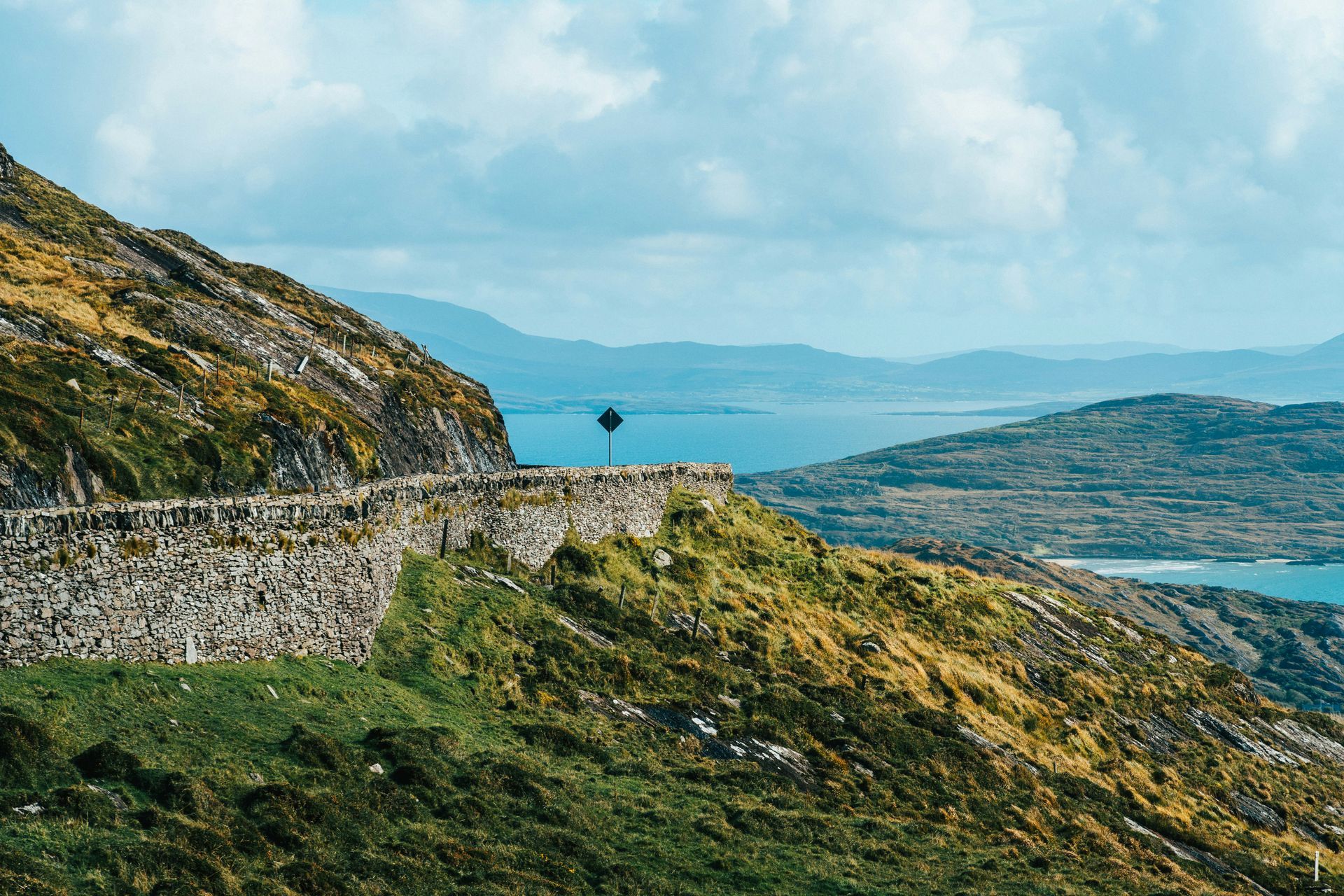 Winding stone road on a mountainside overlooking the sea, under a partly cloudy sky on the Ring of Kerry, Ireland.