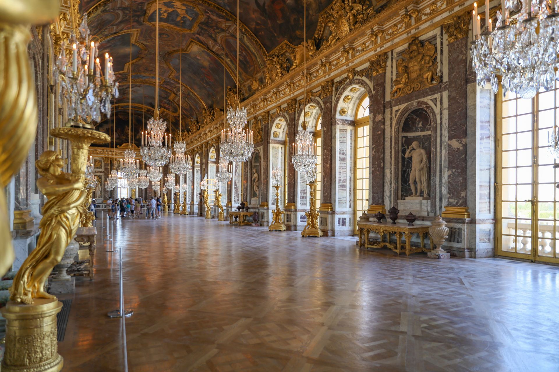 A large room with a lot of windows and statues in it at the Palace of Versailles in France.