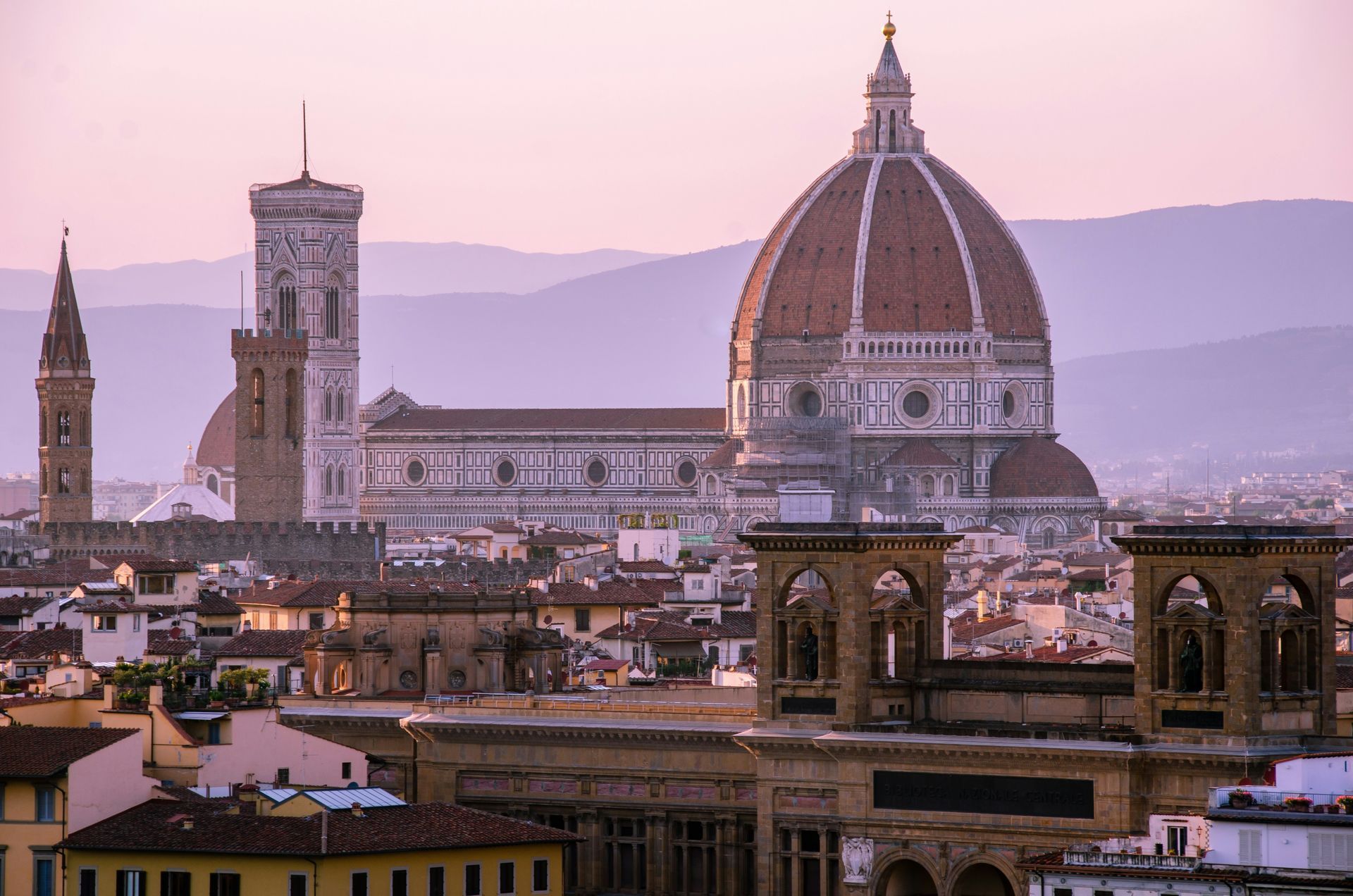 An aerial view of Florence with the Cathedral of Santa Maria del Fiore in the foreground and mountains in the background.