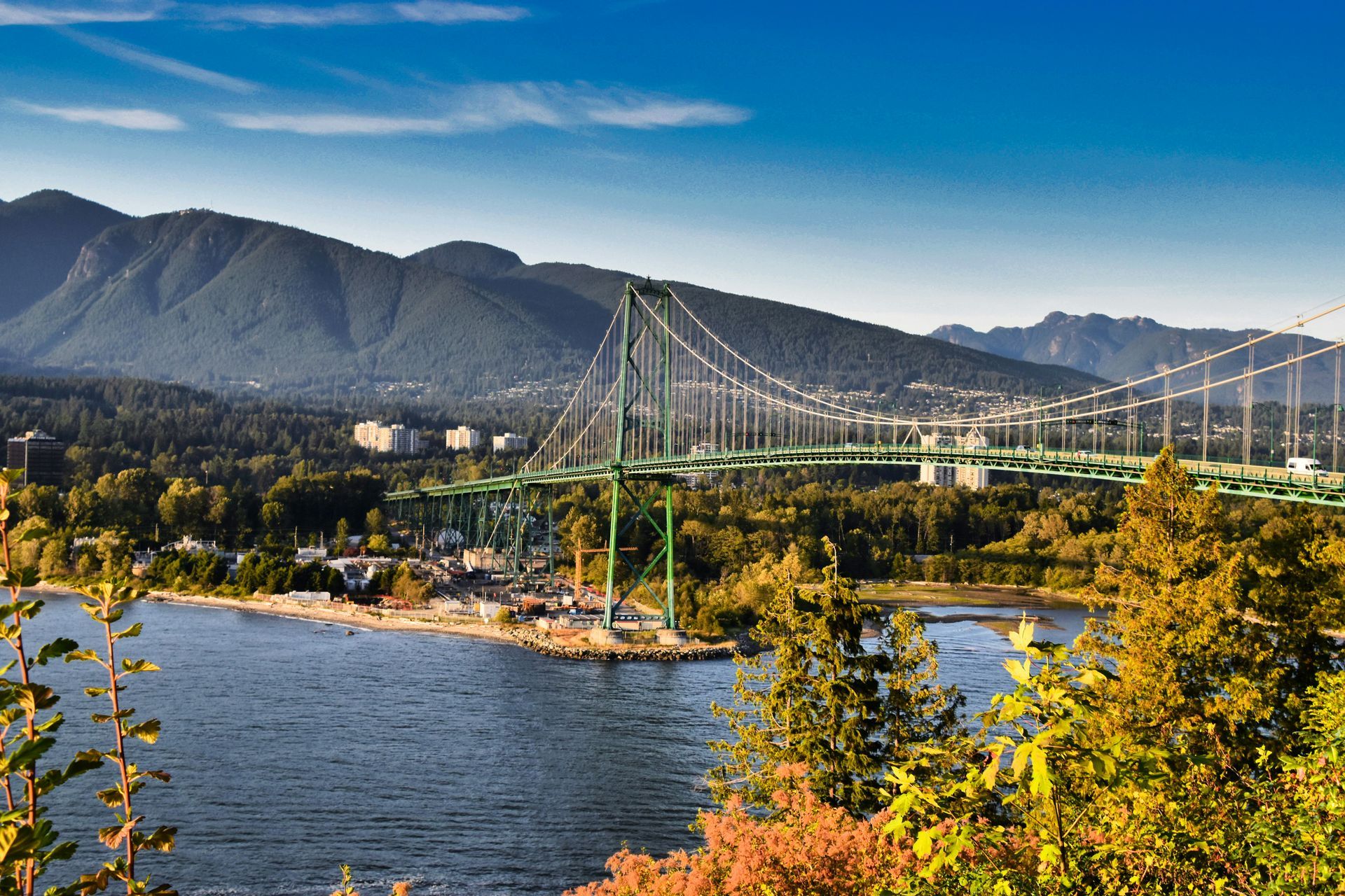 Lions Gate Bridge in Vancouver, BC, spanning blue water with forested mountains in the background under a bright blue sky.