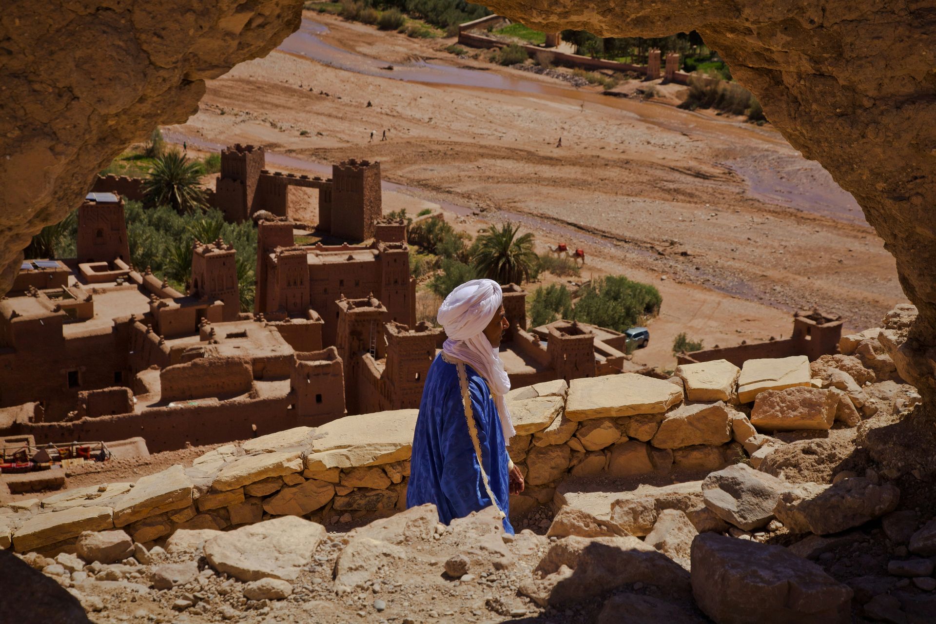 A man in a turban is standing on top of a rocky hill overlooking a desert landscape and the town of Merzouga, Morocco.