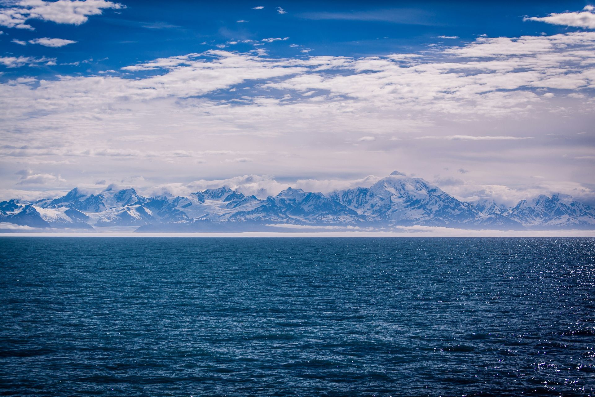 Ocean view with snowy mountains on the horizon, under a partly cloudy blue sky in The Northwest Passage, Canada.