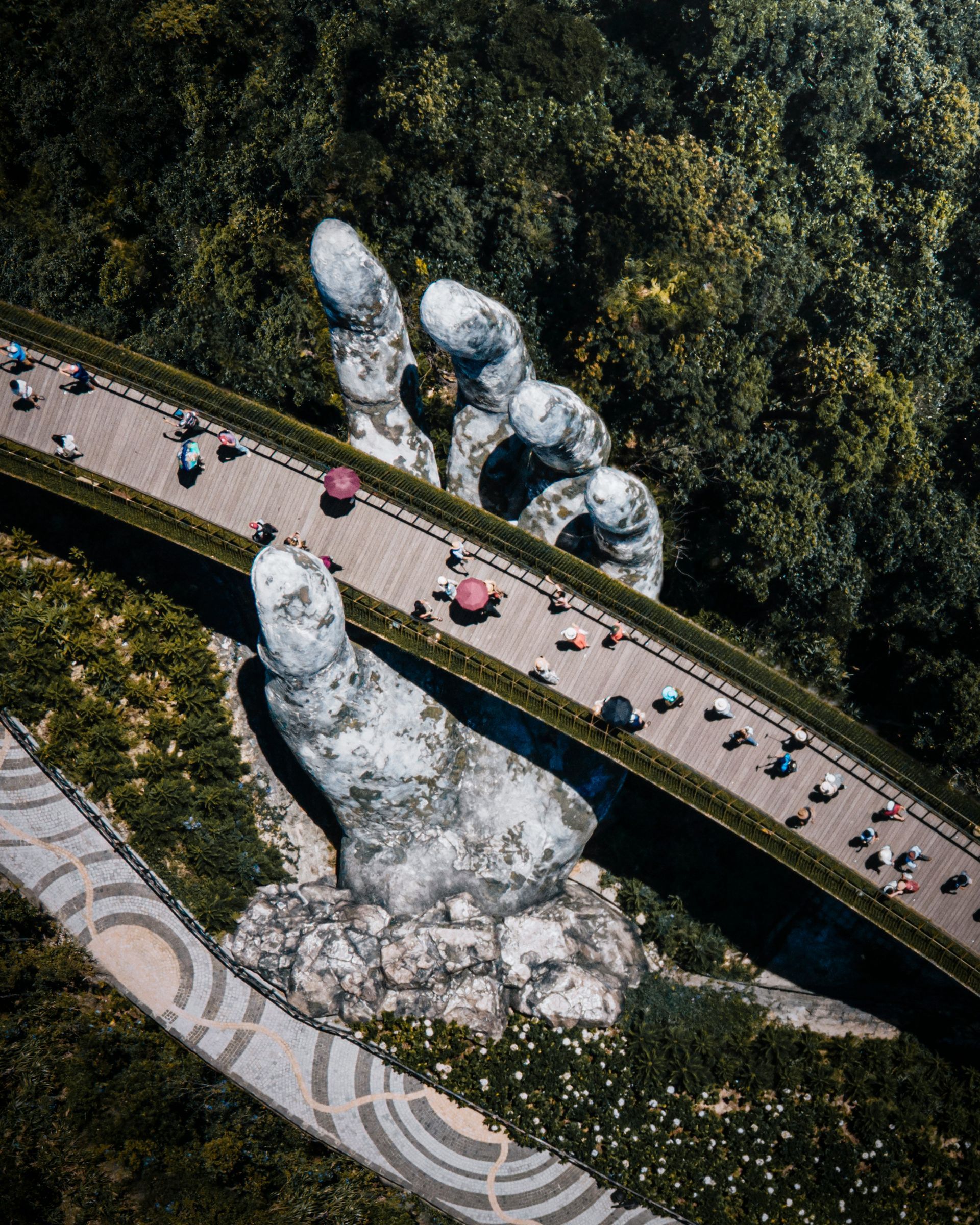 Aerial view of the Golden Bridge in Vietnam, held up by giant stone hands. People walk the path; lush green surroundings.