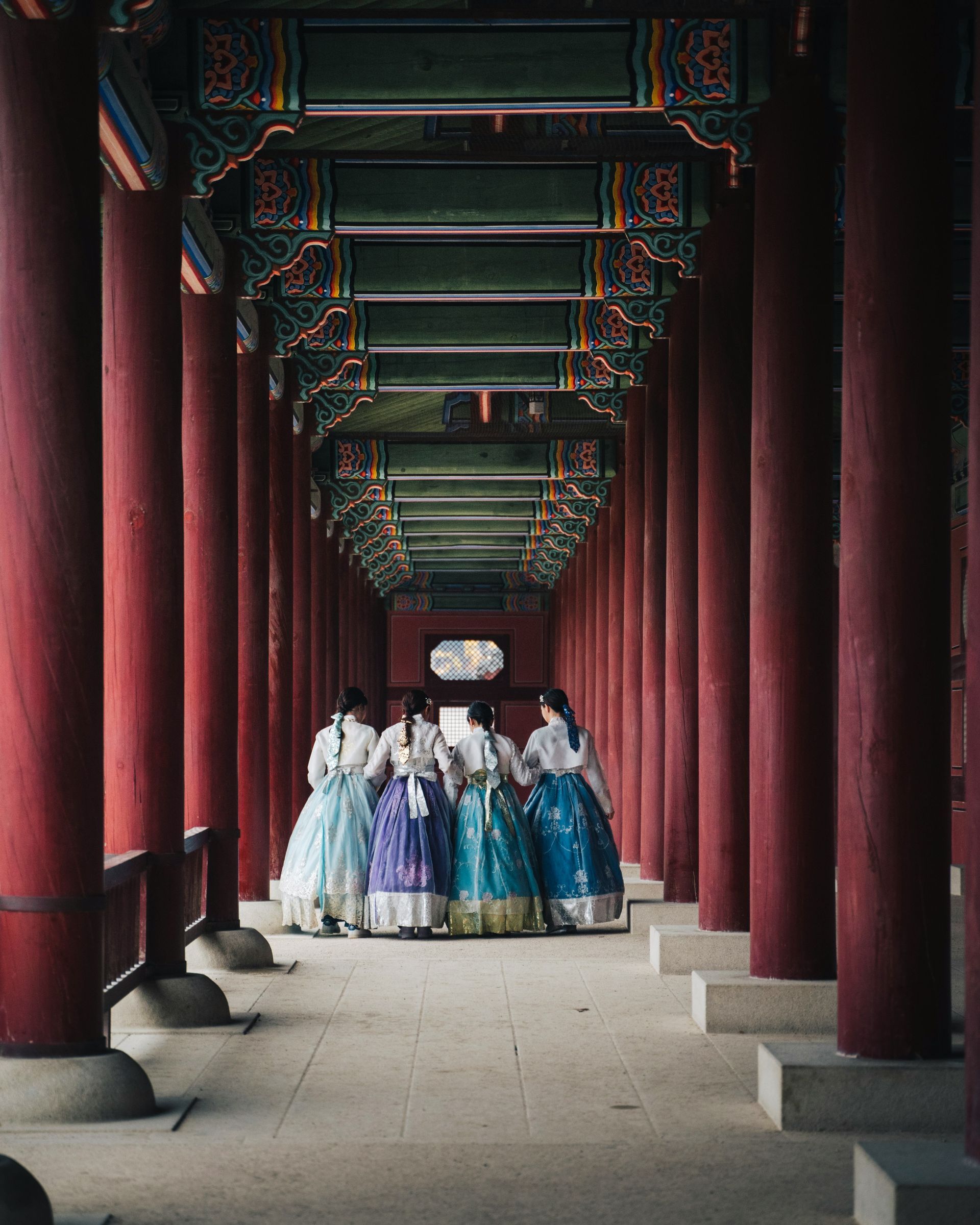 People in traditional Korean hanboks walk down a red-pillared hallway in South Korea.