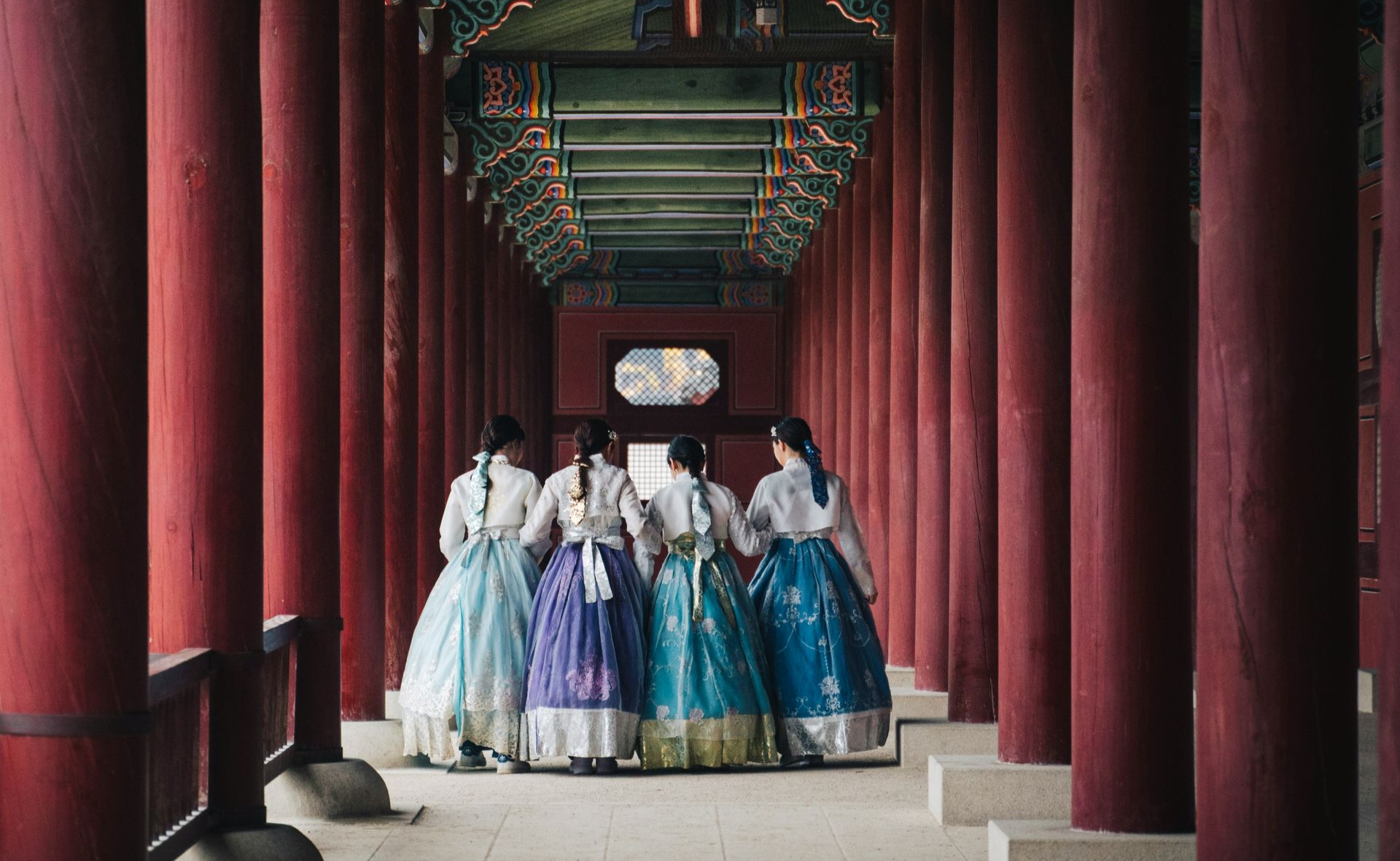 Five women in colorful hanboks walk away through a red-pillared corridor with an ornate ceiling in South Korea.